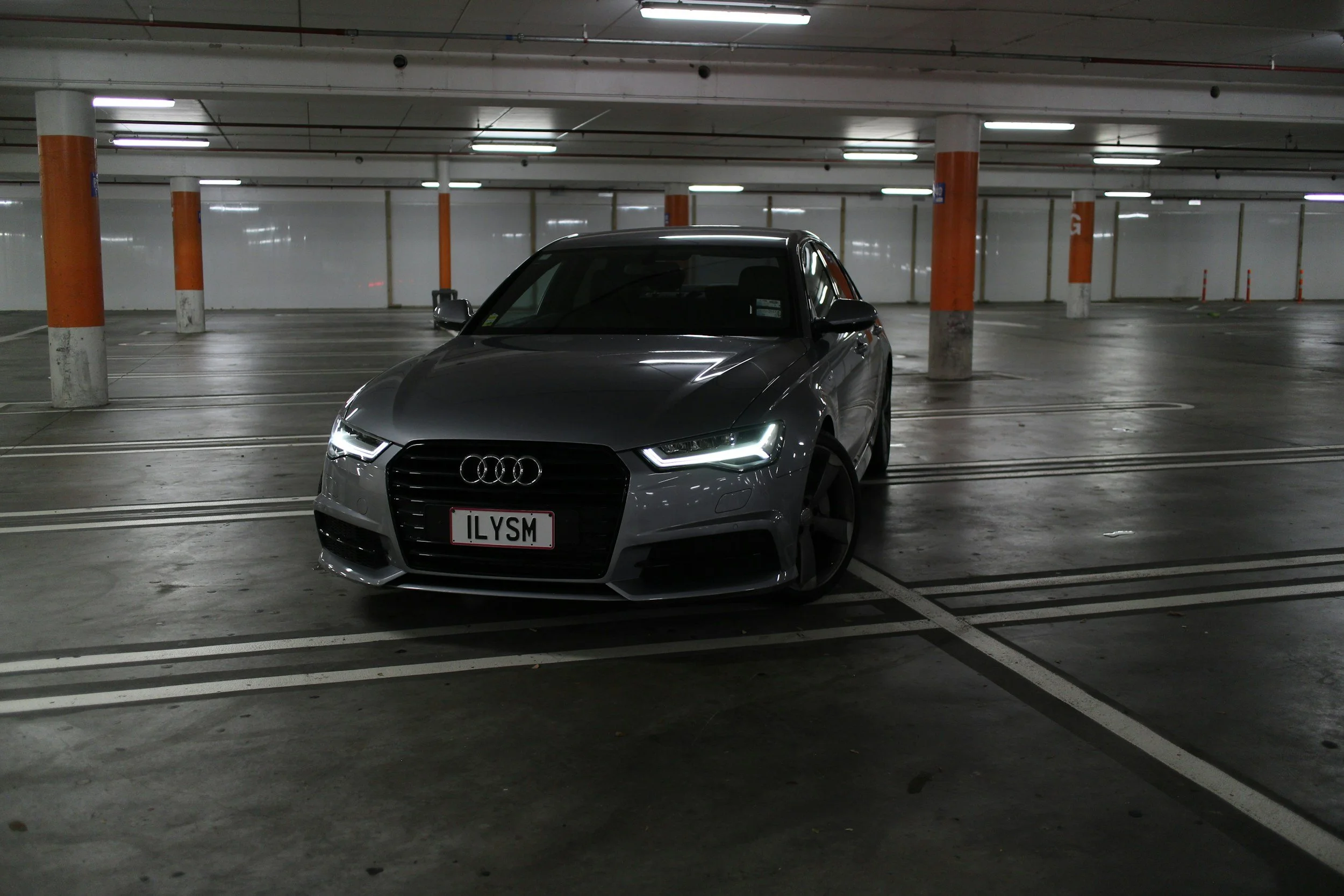 Gray Audi sedan parked in a mostly empty underground parking garage with orange and white pillars and fluorescent lighting