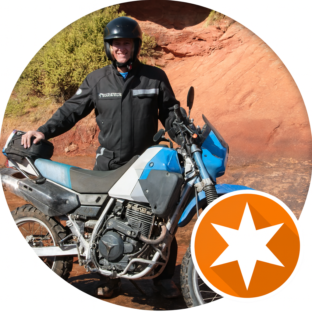 A person wearing a black helmet and jacket standing next to a blue and white off-road motorcycle in a desert landscape with red rocks and sparse green shrubbery.