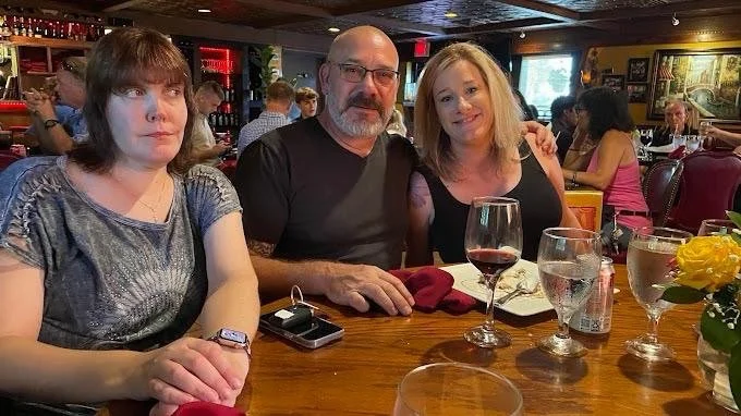 Three people sitting at a restaurant table with drinks and flowers, posing for a photo.
