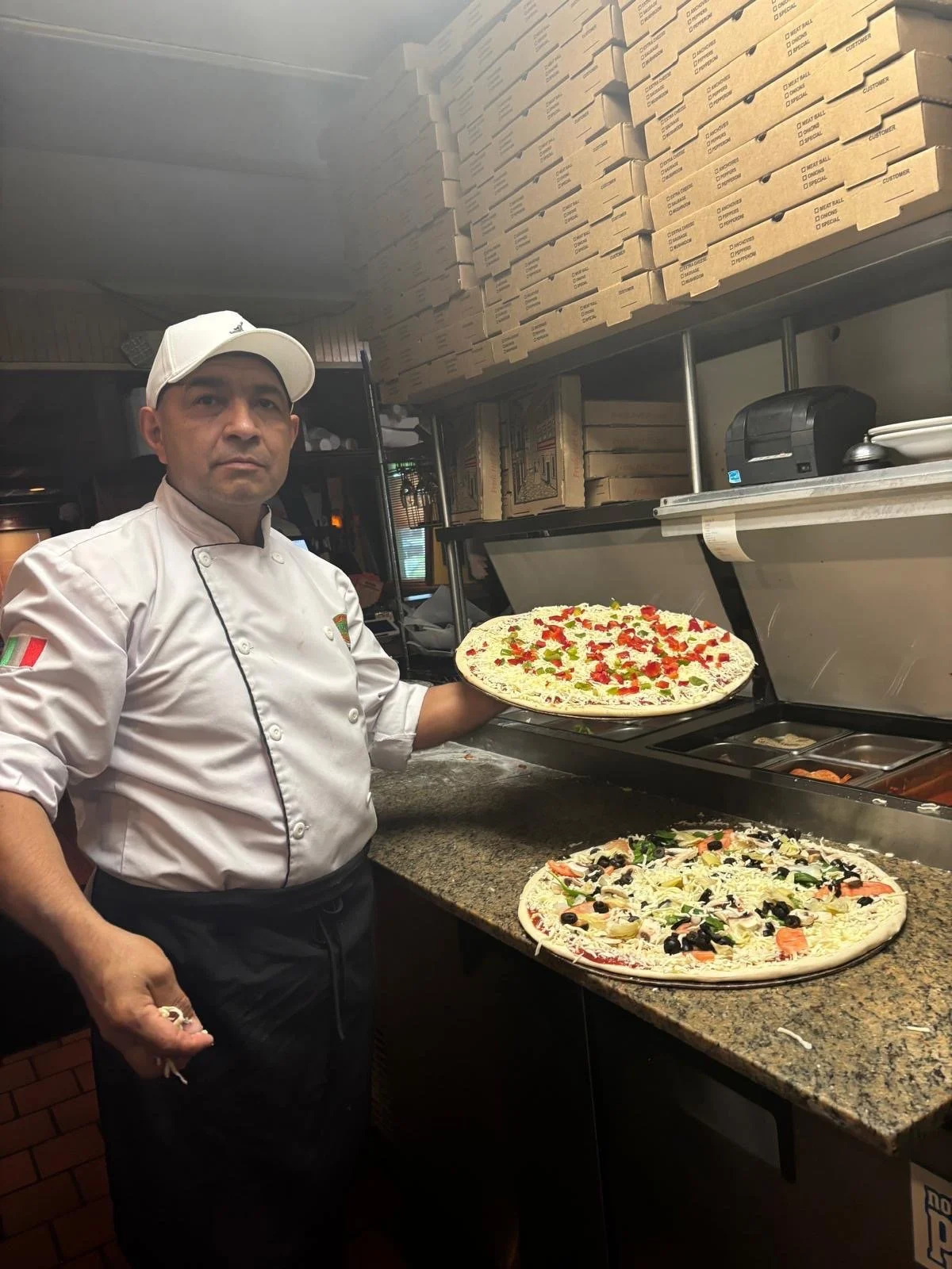A chef in a white uniform and hat stands in a pizzeria kitchen, holding a pizza with toppings of red peppers, green herbs, and cheese, while another pizza with black olives, vegetables, and cheese rests on the counter.