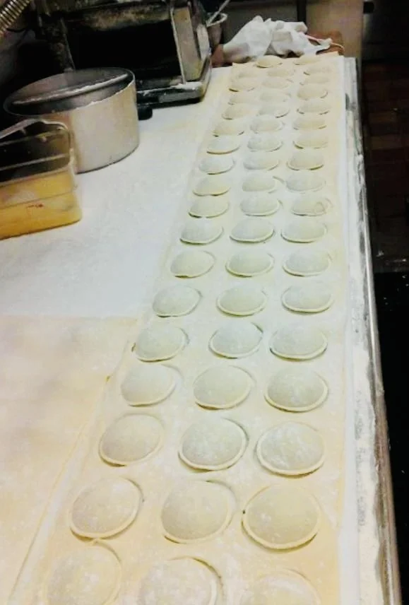 Rows of uncooked dumplings or ravioli shaped pasta on a floured surface in a kitchen.