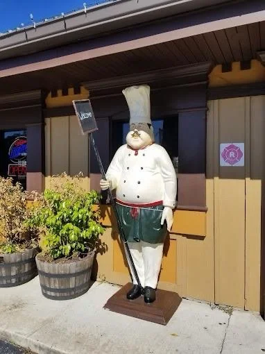 Plastic chef statue holding a sign outside a restaurant with potted plants nearby.