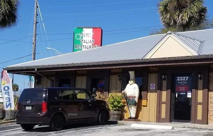 Exterior of a restaurant with a life-sized ceramic chef statue and a black vehicle parked in front.
