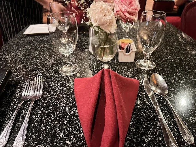 A restaurant table set for two with a black speckled granite surface, featuring a red folded napkin in the center, two empty wine glasses, a small floral arrangement with pink and white roses, and a container with sugar packets.