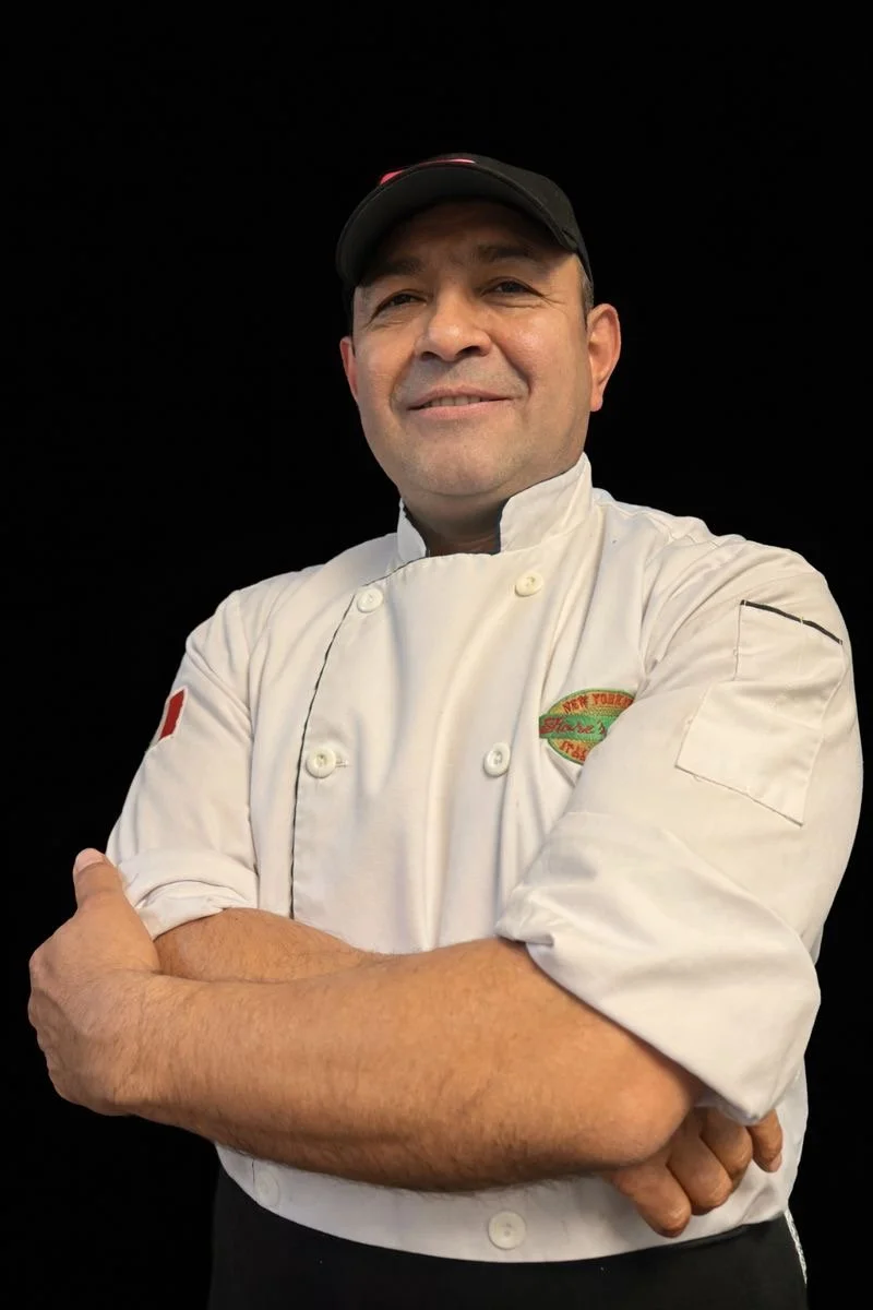 A male chef in a white chef coat and black cap standing with arms crossed, smiling, against a black background.