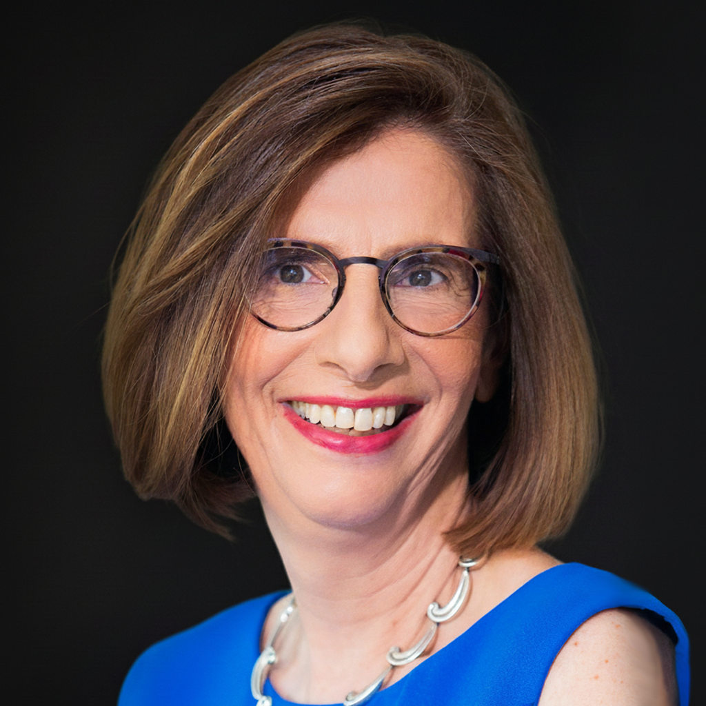 A woman with shoulder-length brown hair, wearing glasses, a blue top, and a silver necklace, smiling against a black background.