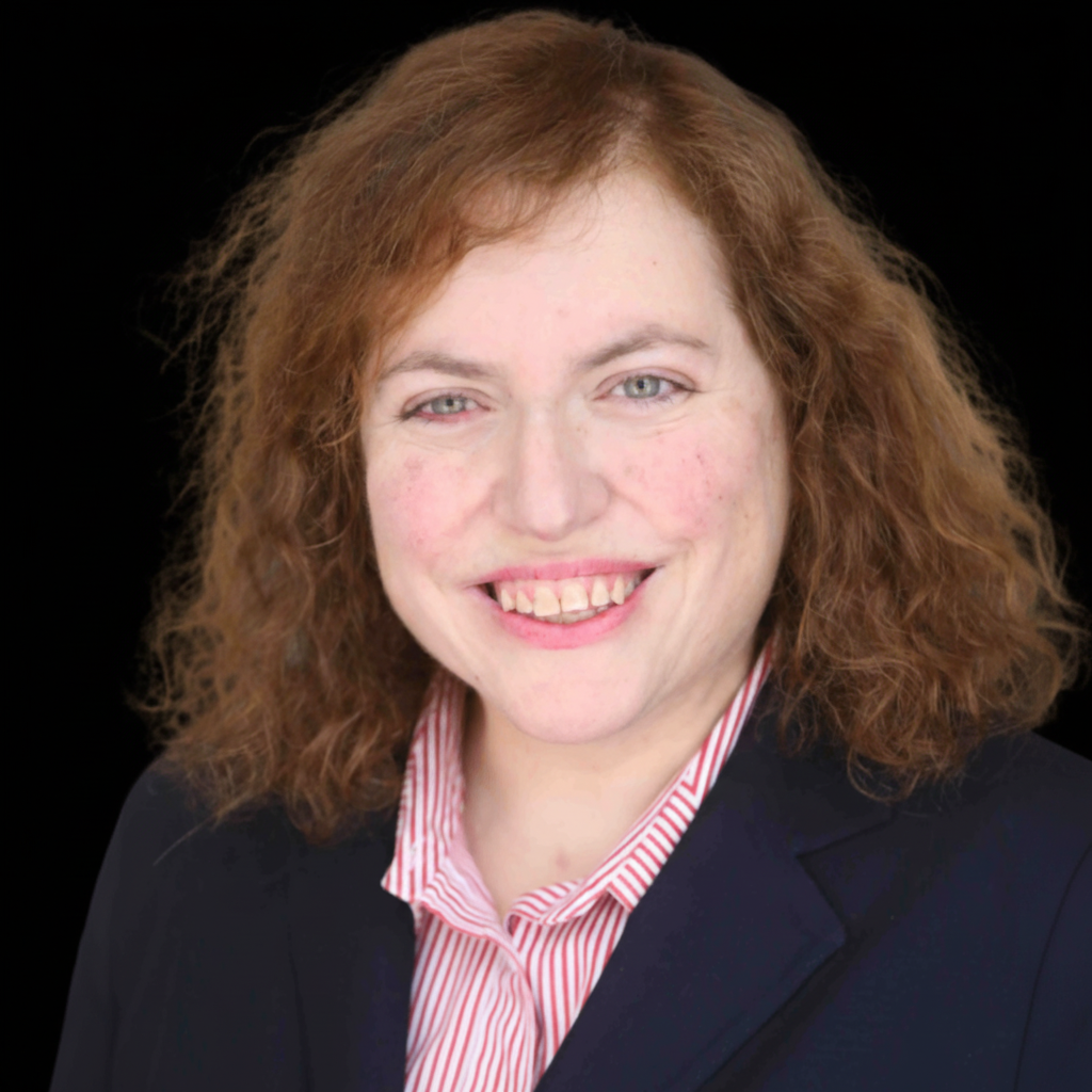 A woman with red curly hair smiling, wearing a dark blazer and a red and white striped shirt, against a black background.