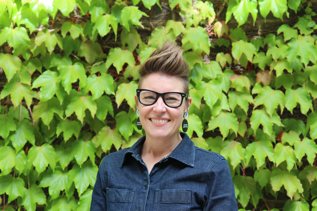 A person with short hair standing in front of a leafy wall. They are wearing glasses and smiling at the camera.