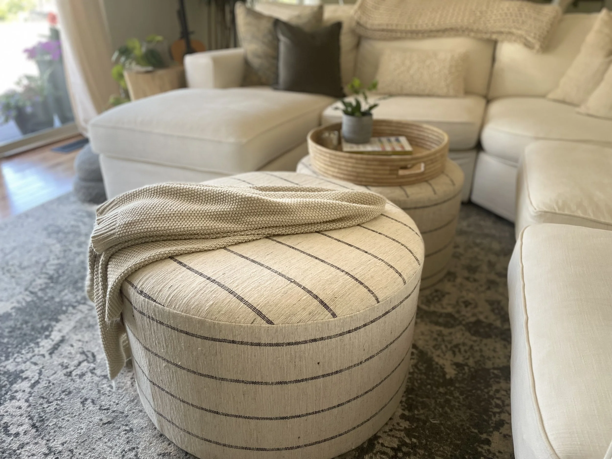 Living room with beige and white sectional sofa, round ottoman with striped fabric, blanket draped over ottoman, and a tray with a plant and magazines on a small round table. Glass door with balcony and plants outside