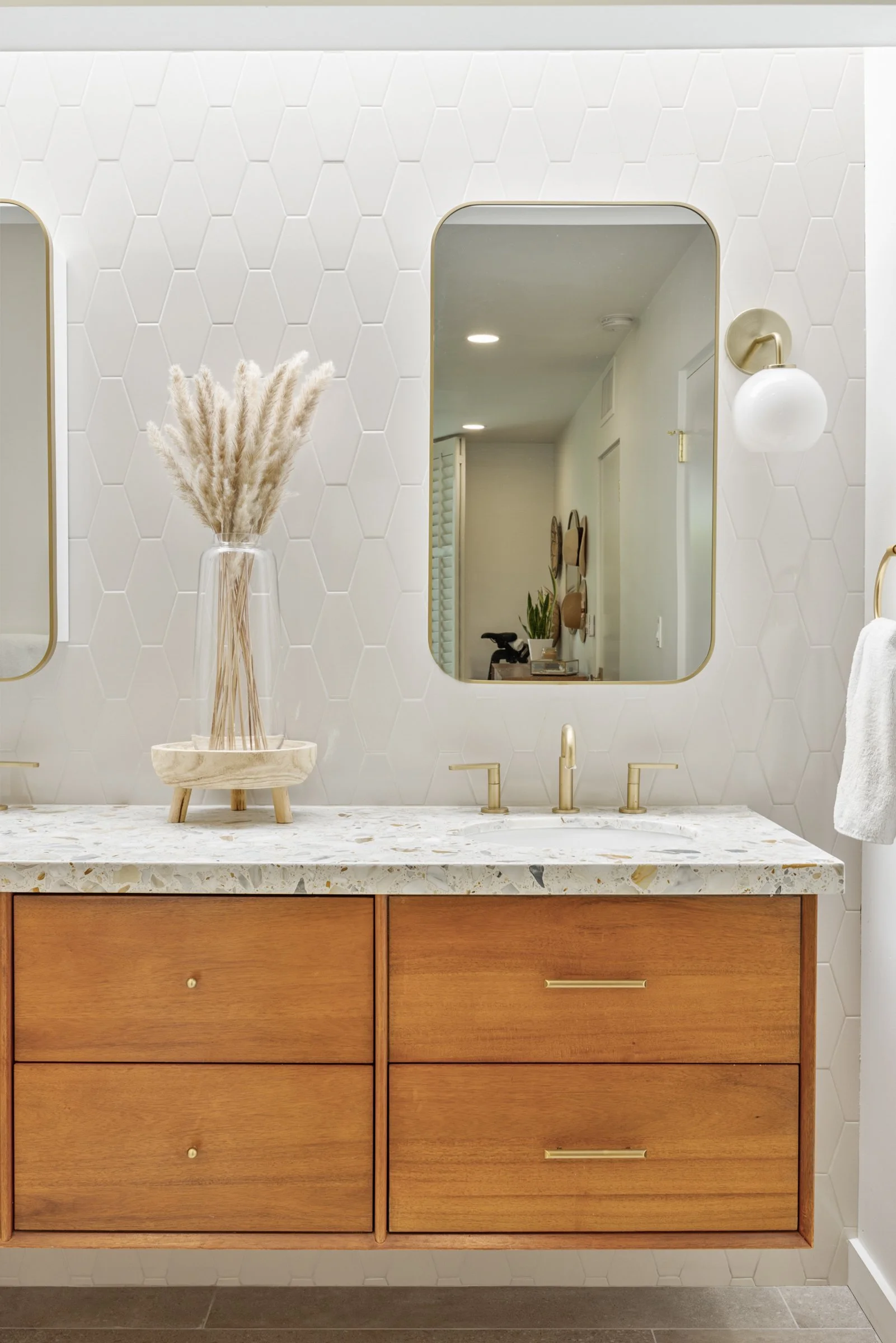 Minimalist bathroom vanity with a wooden cabinet, marble countertop, gold faucet, white hexagonal tiled wall, mirror, and decorative vase with pampas grass.