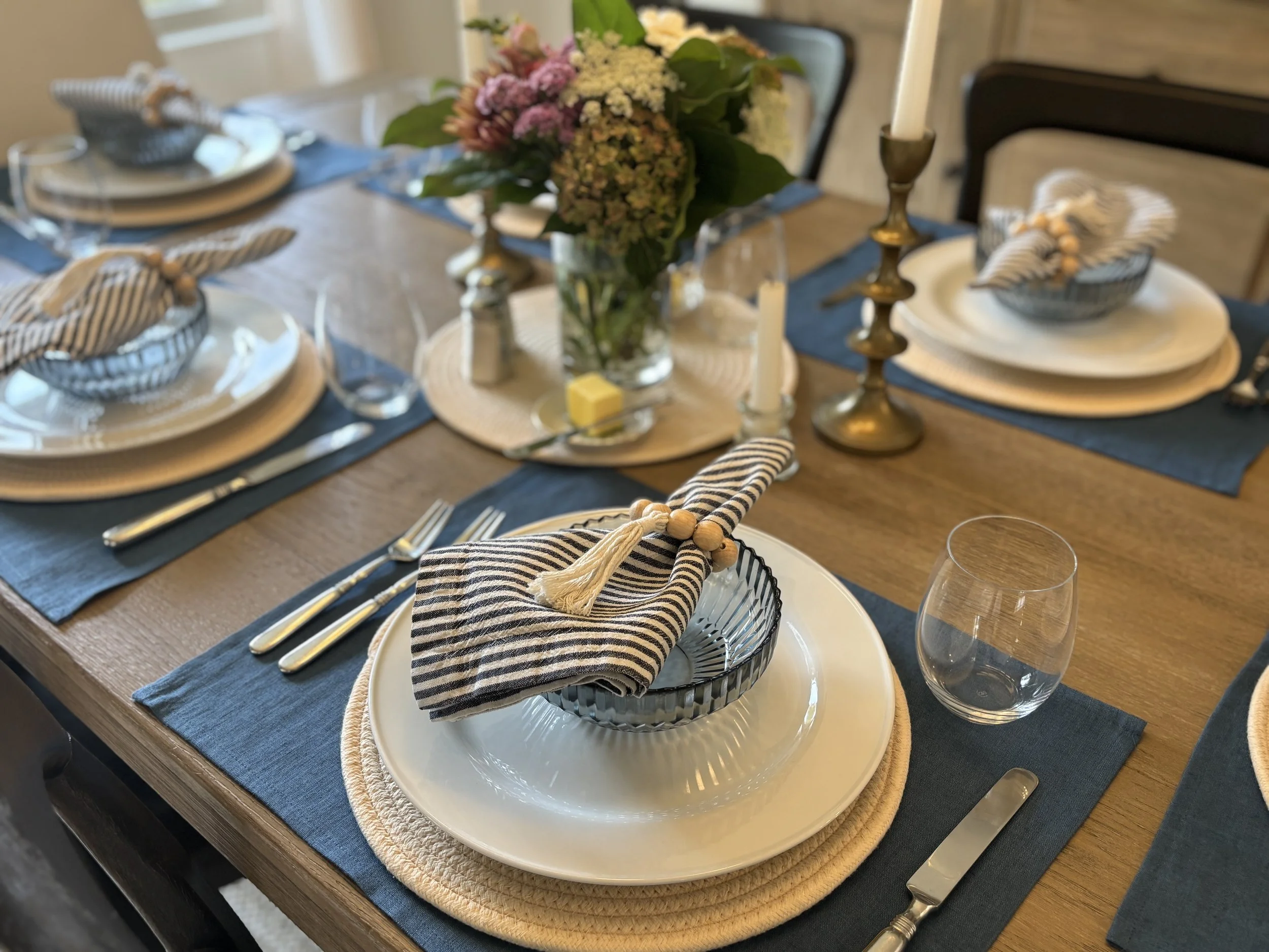 A dining table set with plates, silverware, cloth napkins in striped patterns, and glassware. There are floral centerpieces and candle holders as decorations.