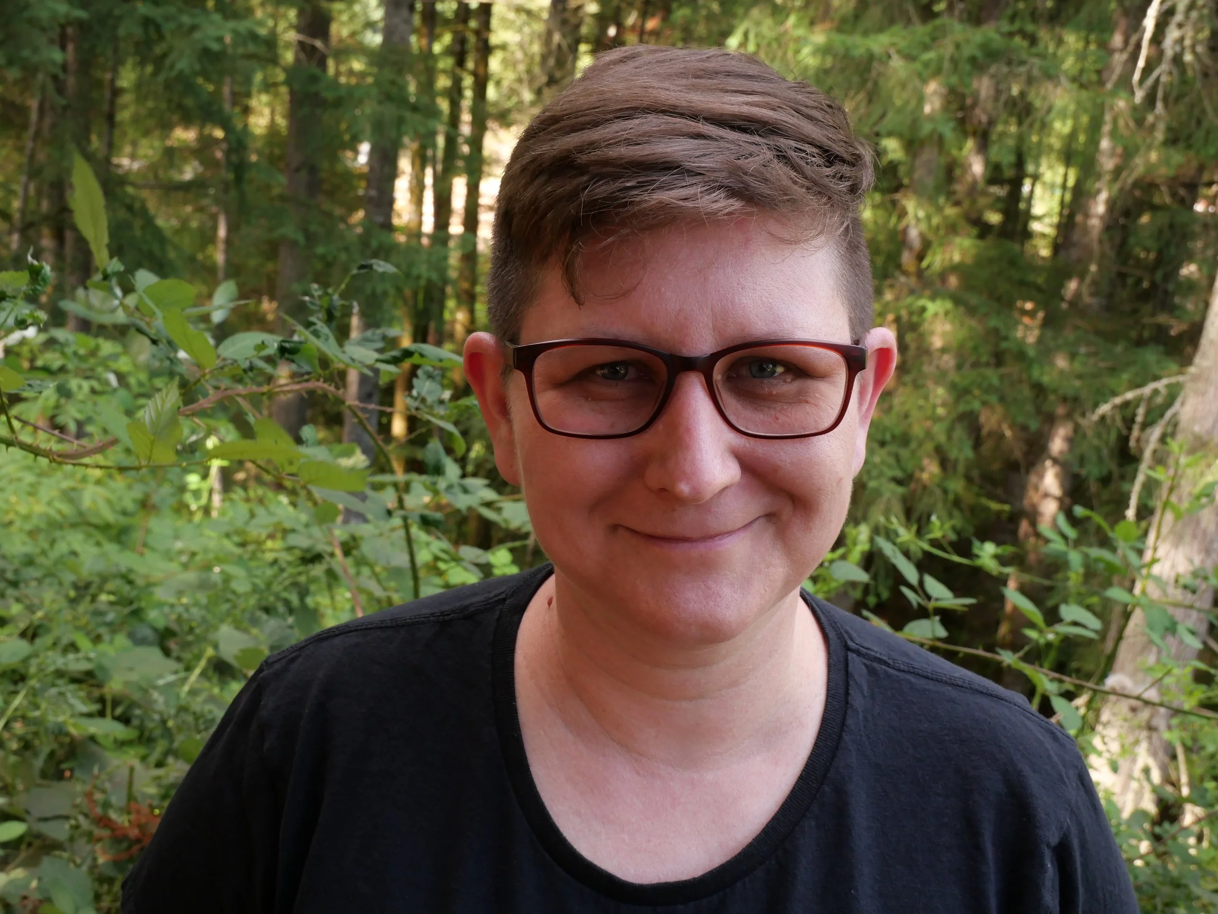 A person with short brown hair, wearing glasses and a black shirt, smiling outdoors in a wooded area with green foliage in the background.