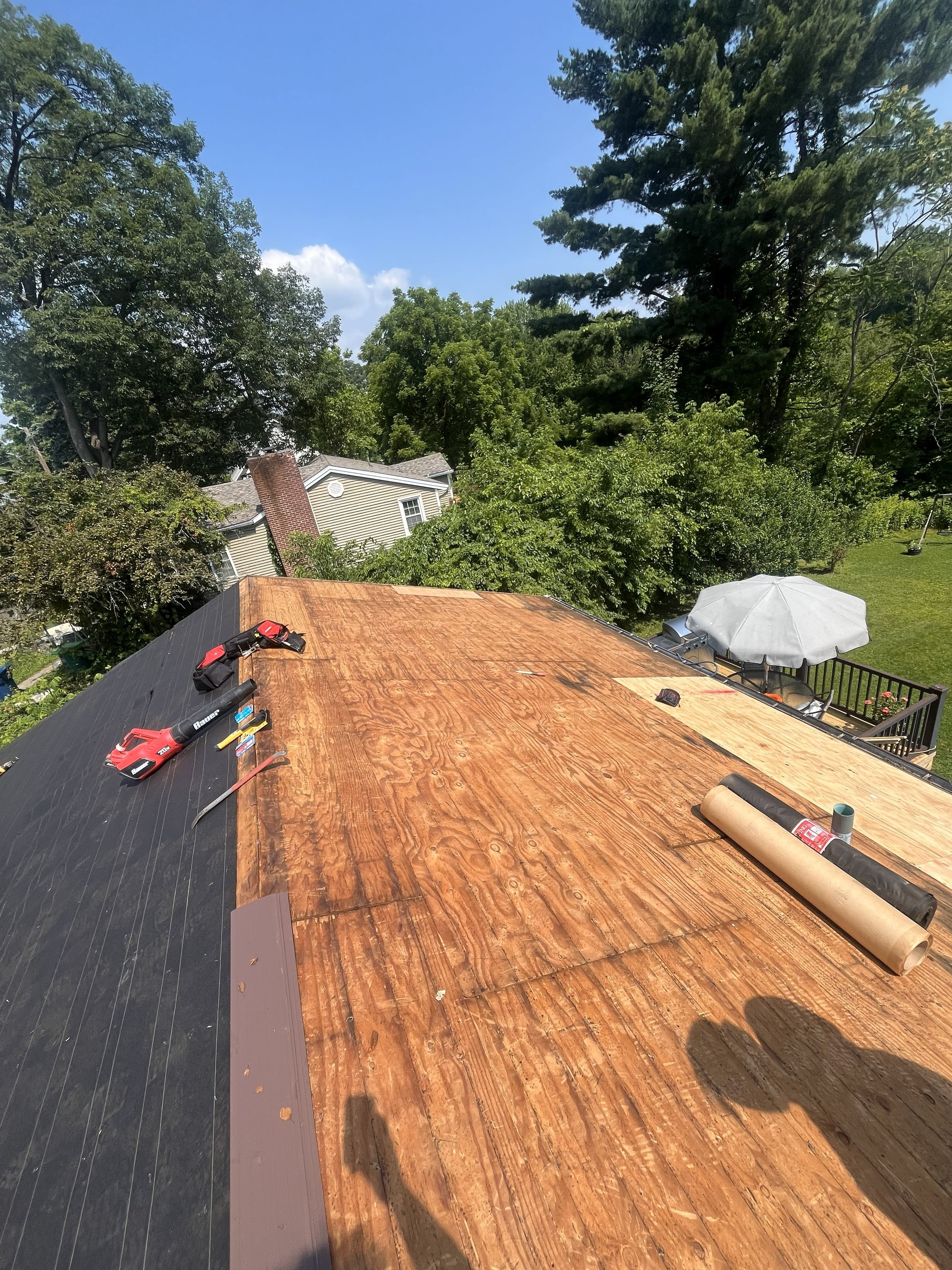 Roof being renovated with new plywood installed, tools and materials on the roof, house, trees, and backyard visible in the background.