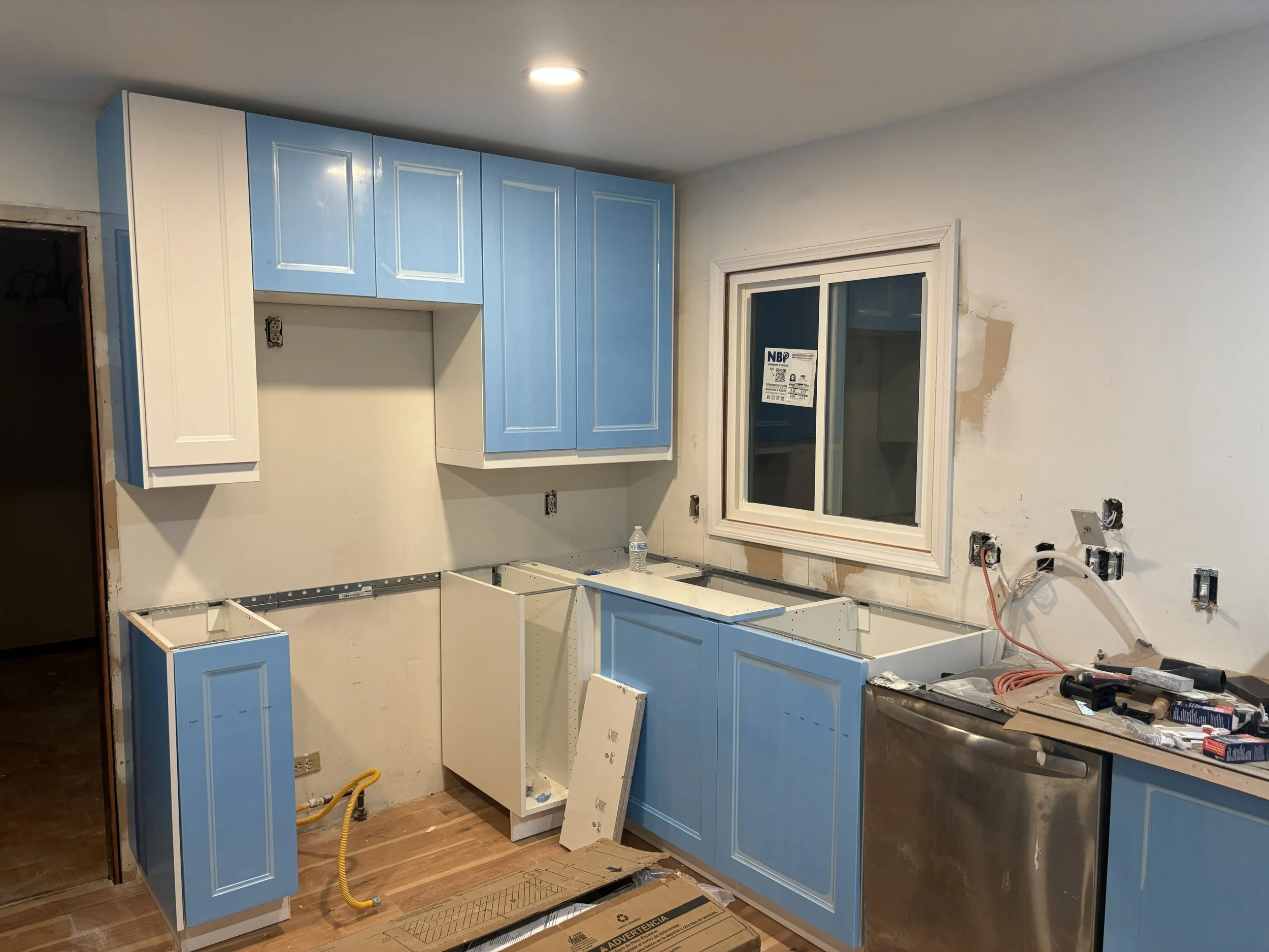 Kitchen under renovation with light blue cabinets, some missing doors, a window, and construction tools on the countertop.