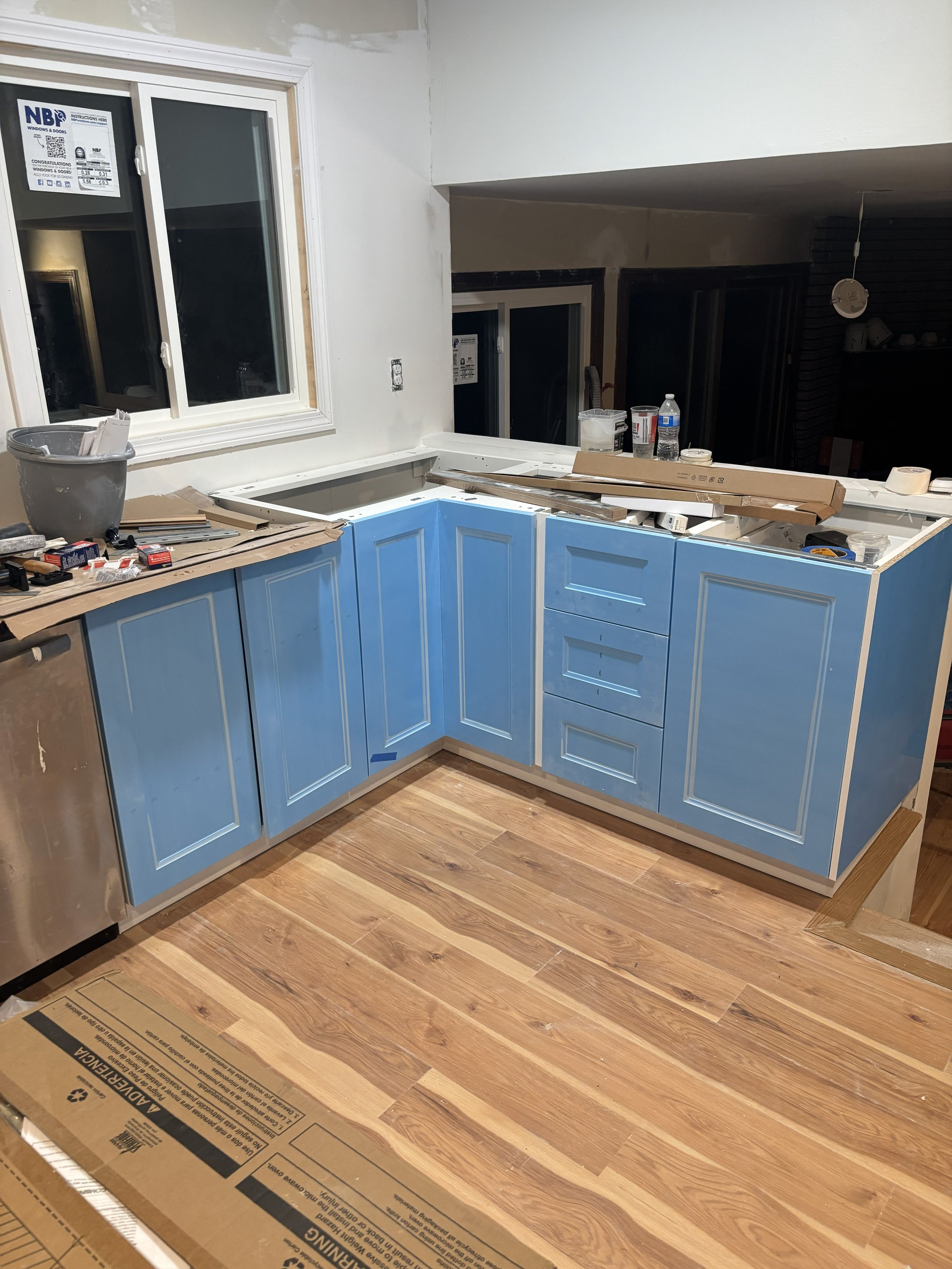 Kitchen under renovation with blue cabinets, a window, and construction materials on the counter and floor.