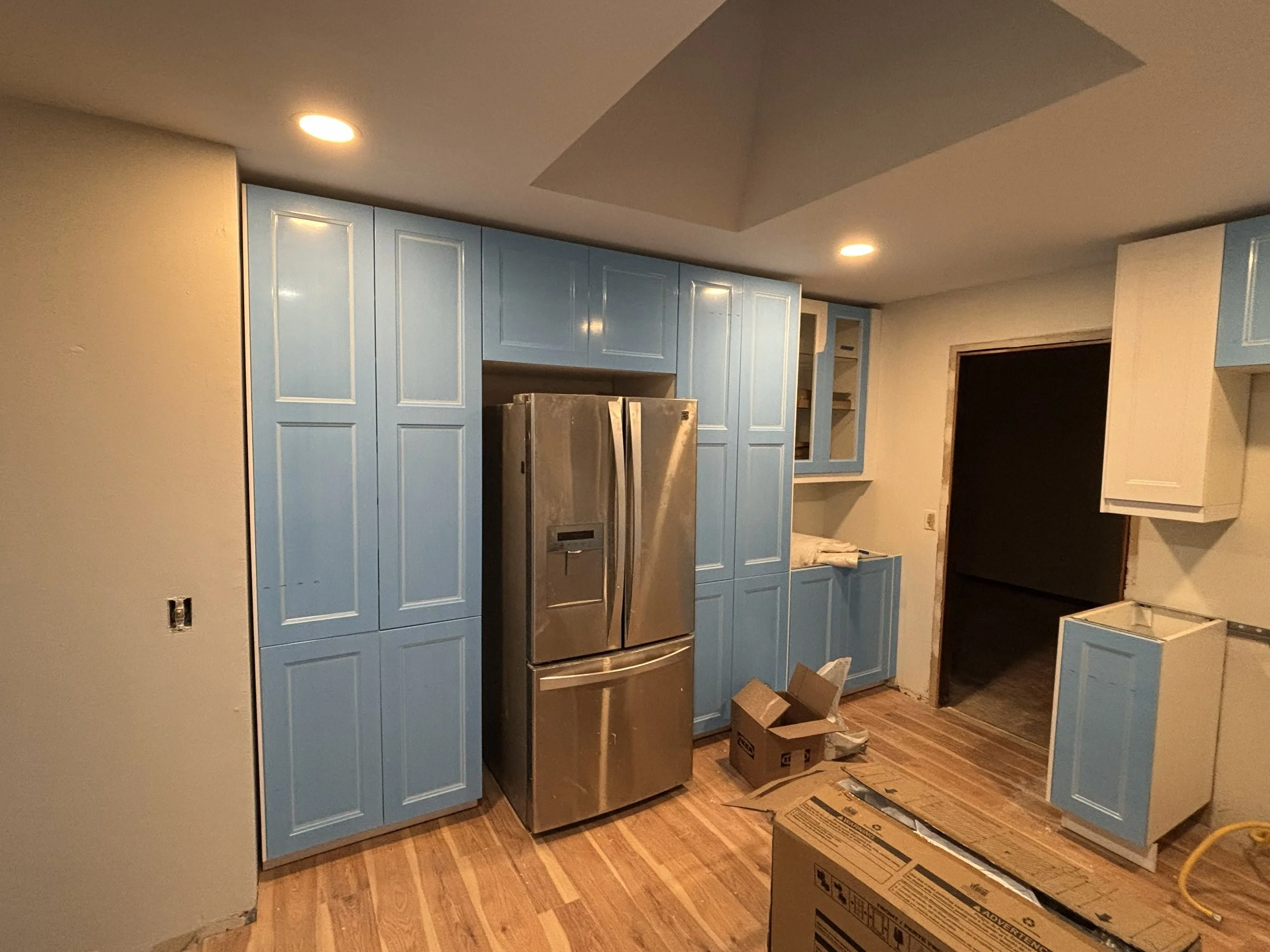 Kitchen under construction with blue cabinets, stainless steel refrigerator, hardwood floors, and an empty space for appliances.