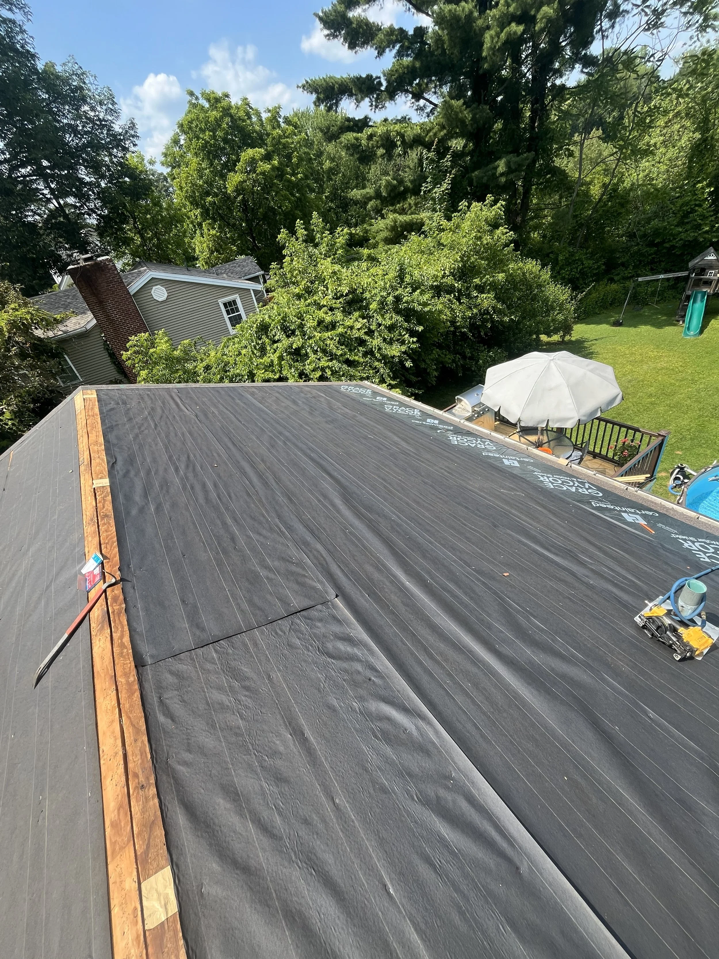 Unfinished roof with black underlayment, a roll of roofing material, a utility knife, and a pipe on the rooftop. In the backyard, there is a patio with an umbrella, a swing set, and a slide. The sky is clear with some clouds, and trees and neighboring houses are visible.
