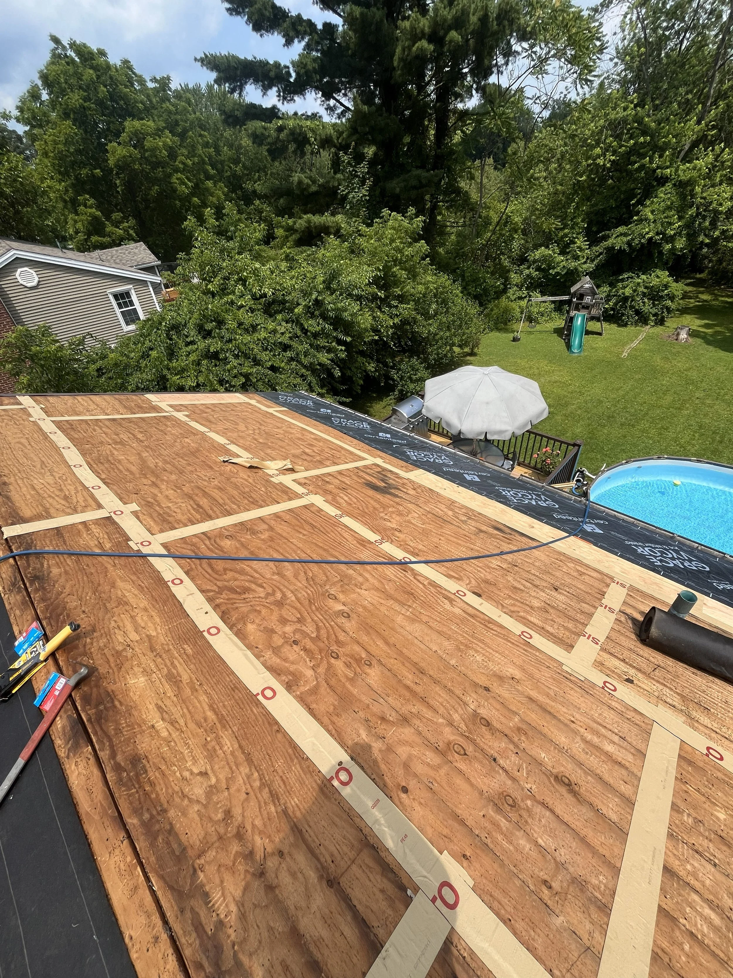 Roof under construction with plywood sheathing and felt paper, overlooking backyard with trees, a shed, a swing set, and a swimming pool.