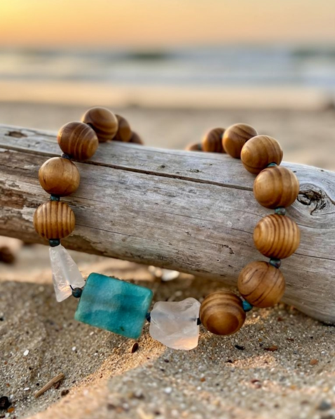 A beaded necklace with wooden beads and large sea glass pendants, resting on a piece of driftwood on a sandy beach at sunset.