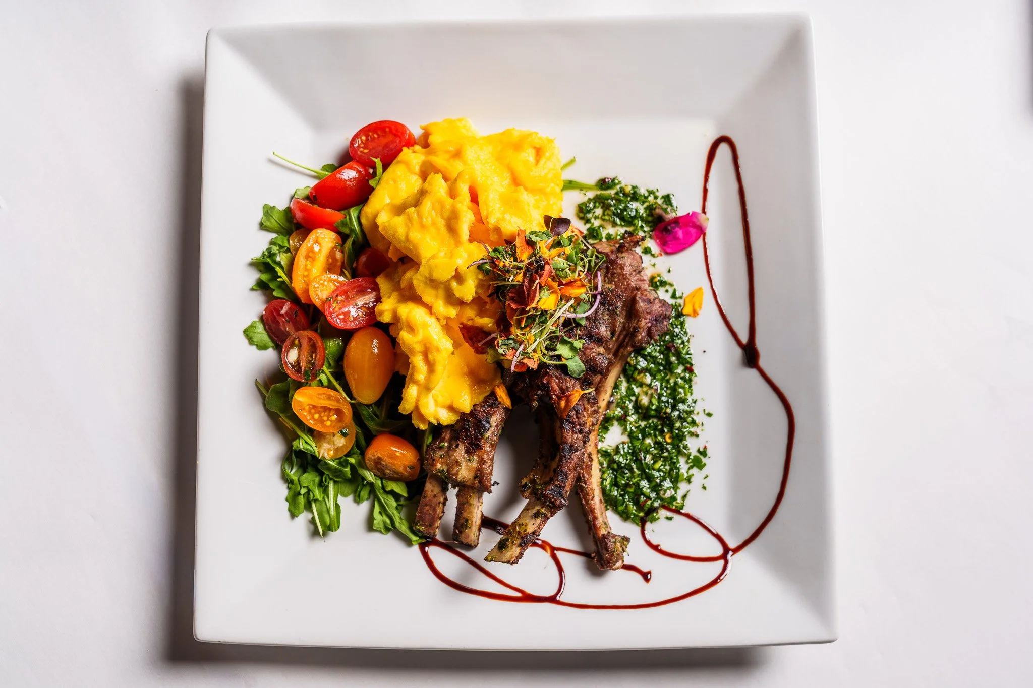 Plate with scrambled eggs, sautéed cherry tomatoes, mixed greens, grilled lamb chops, and garnished with herbs and a drizzle of sauce.