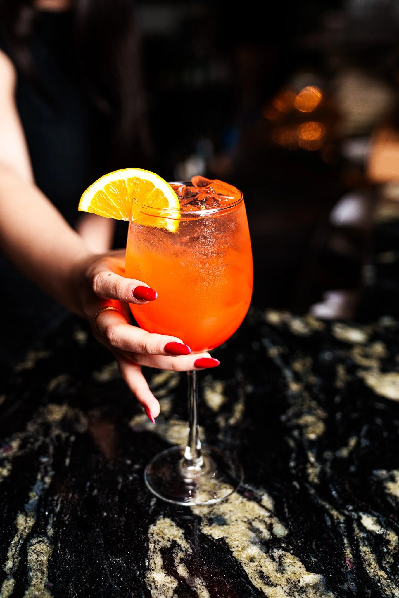 A hand with red painted nails is holding a tall wine glass filled with an orange-colored cocktail and ice cubes, garnished with a slice of orange.