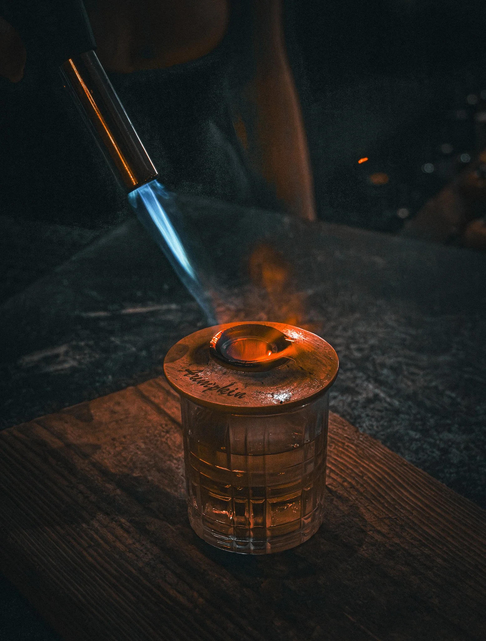 Glass of whiskey on a wooden surface with a metal lid labeled 'Thunderkin' and a blowtorch torching it.