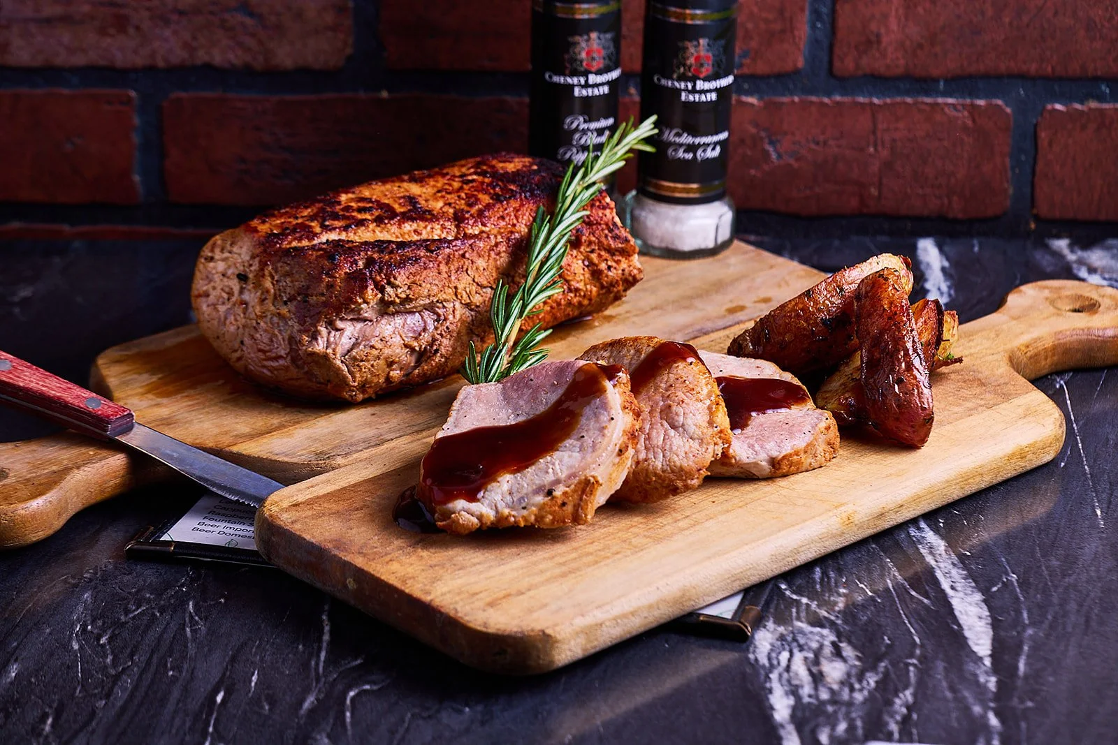 Meatloaf with slices topped with gravy, served on a wooden cutting board with a sprig of rosemary, next to baked potato wedges, on a dark marble surface with salt and pepper shakers in the background.
