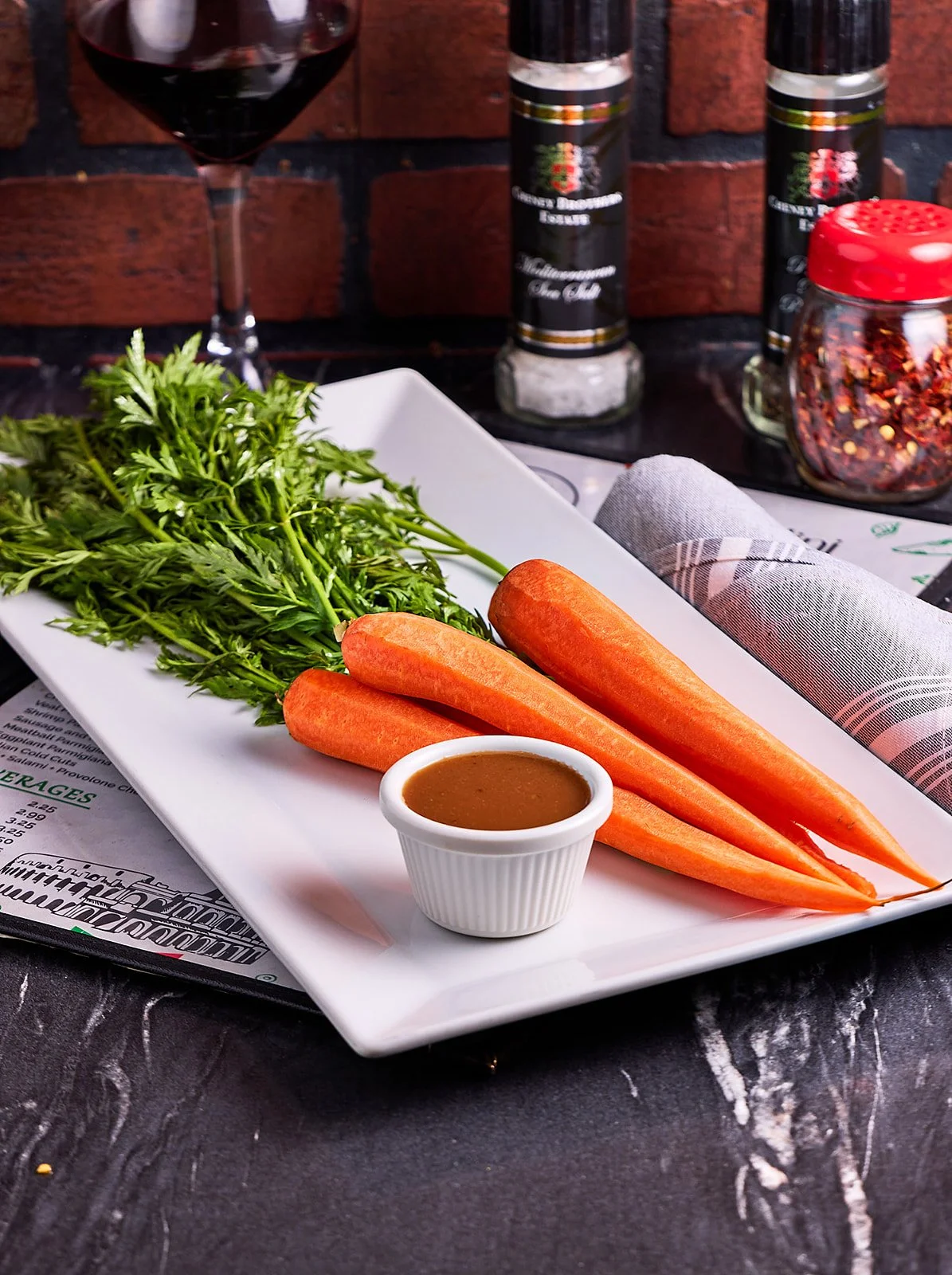 Fresh carrots with green tops on a white rectangular plate, accompanied by a small cup of brown sauce, set on a dark surface with a wine glass, condiment bottles, and a jar of chili flakes in the background.