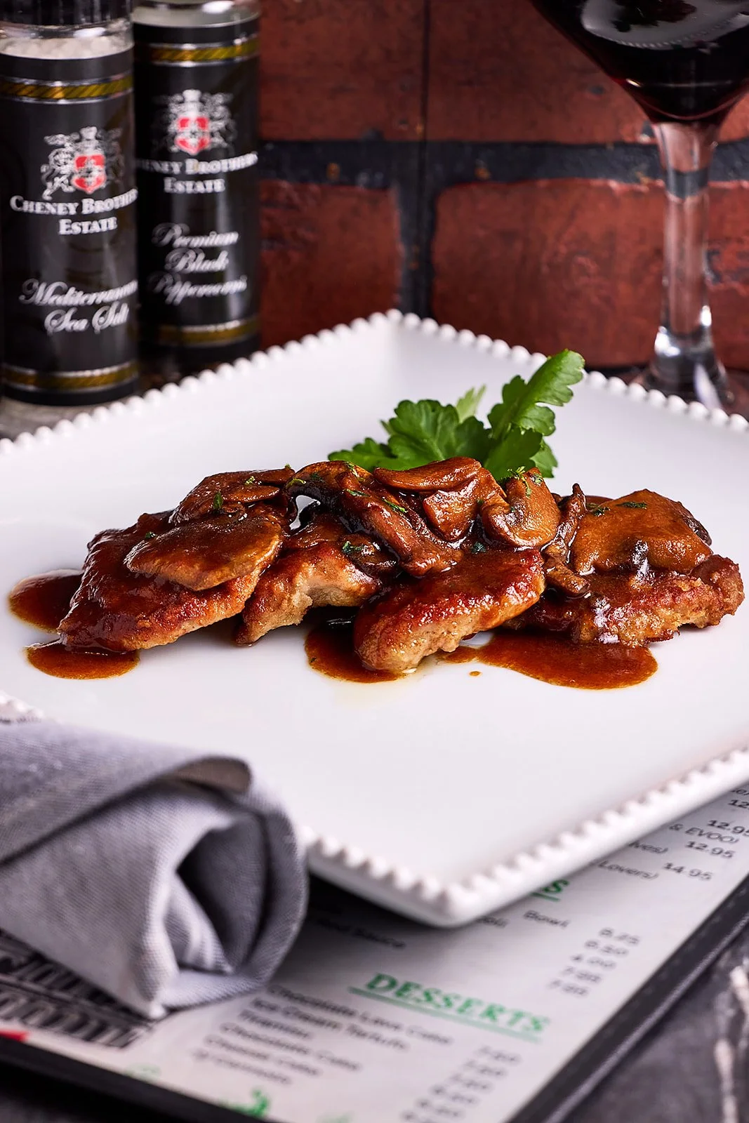 A plate of chicken with mushroom gravy garnished with a sprig of cilantro, and a glass of red wine in the background.