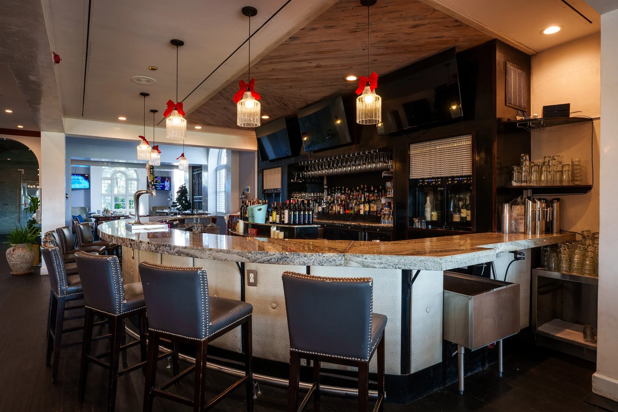 Empty bar with a marble countertop, black leather bar stools, hanging pendant lights with red bows, and a backdrop of liquor bottles and glassware, in a well-lit restaurant or pub setting.