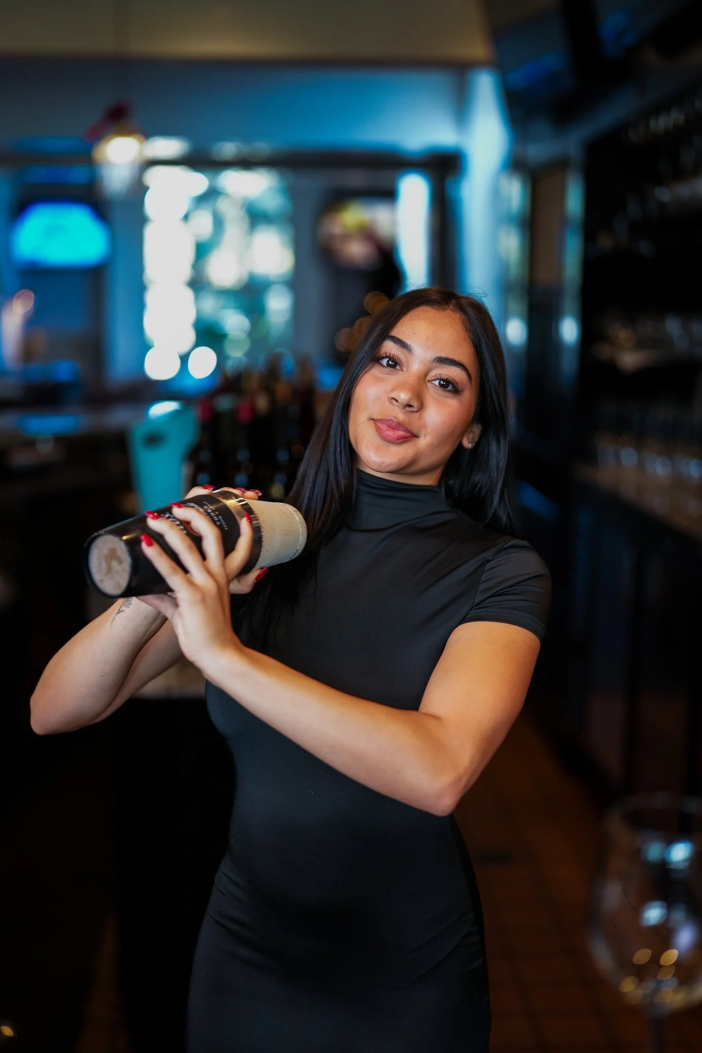 A young woman with long dark hair, wearing a black dress, holding a wine bottle in a restaurant or bar, smiling softly at the camera.