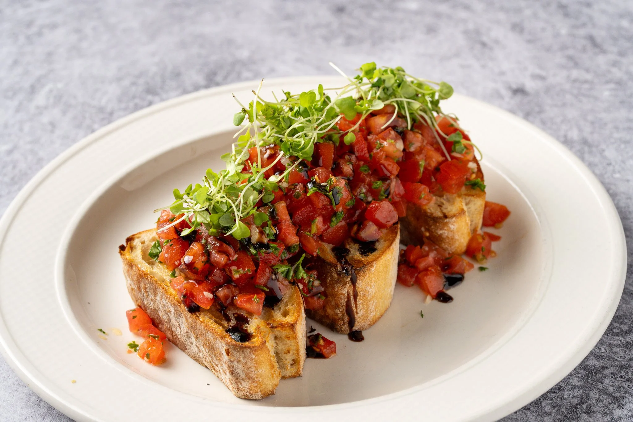 Two pieces of toasted baguette topped with chopped tomatoes, herbs, and microgreens on a white plate.
