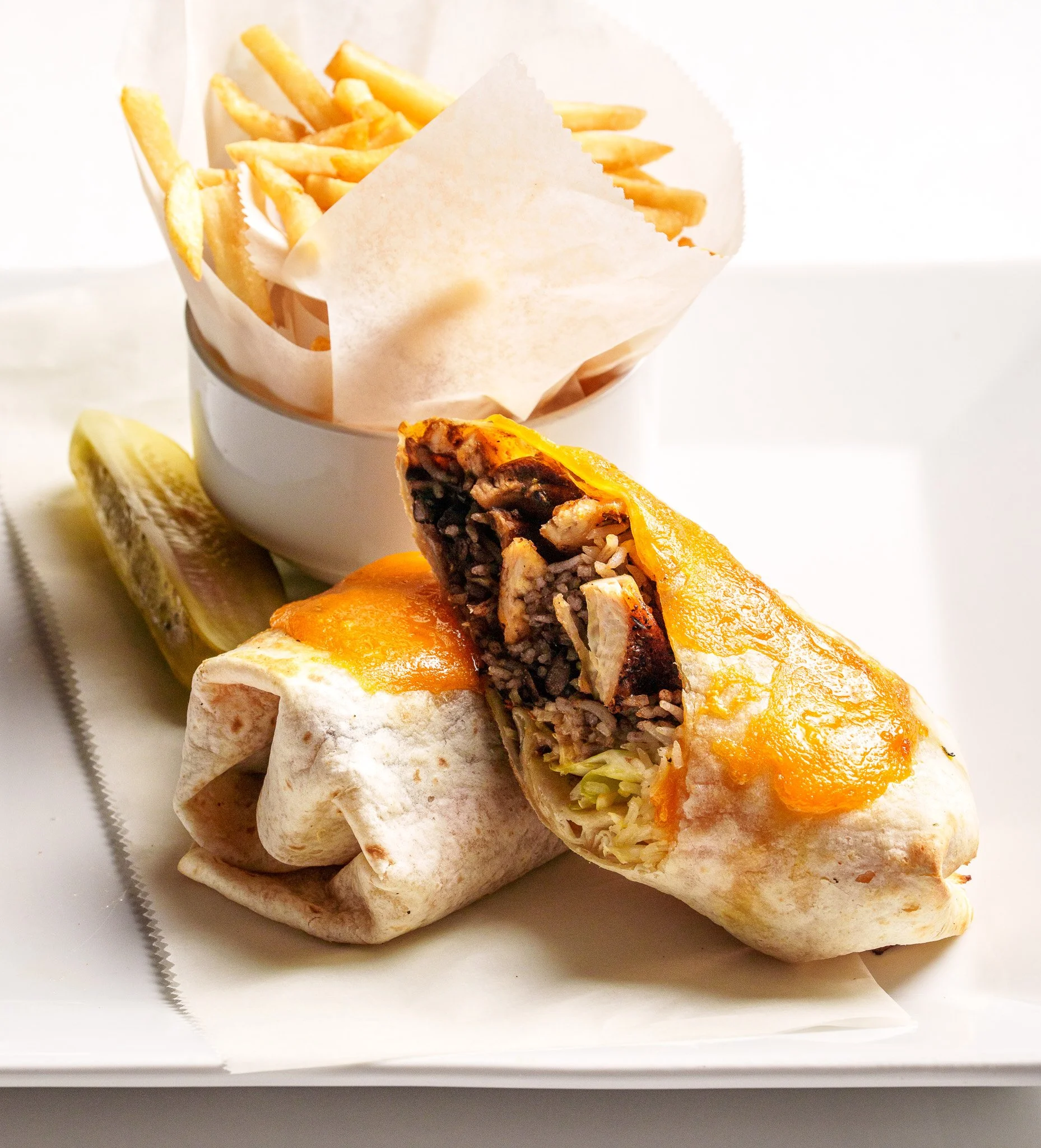 A Mexican burrito with melted cheese, chopped chicken, rice, and beans inside a flour tortilla, accompanied by a pickle spear and a small cup of French fries.