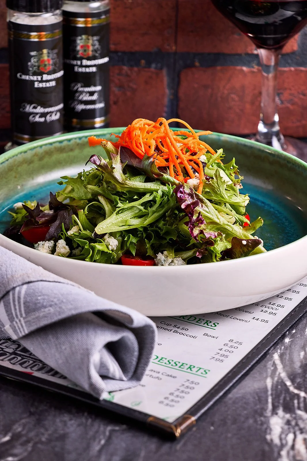 A fresh mixed green salad with shredded carrots, cherry tomatoes, crumbles of cheese, served in a bowl on a restaurant table with a menu underneath, set against a brick wall background.
