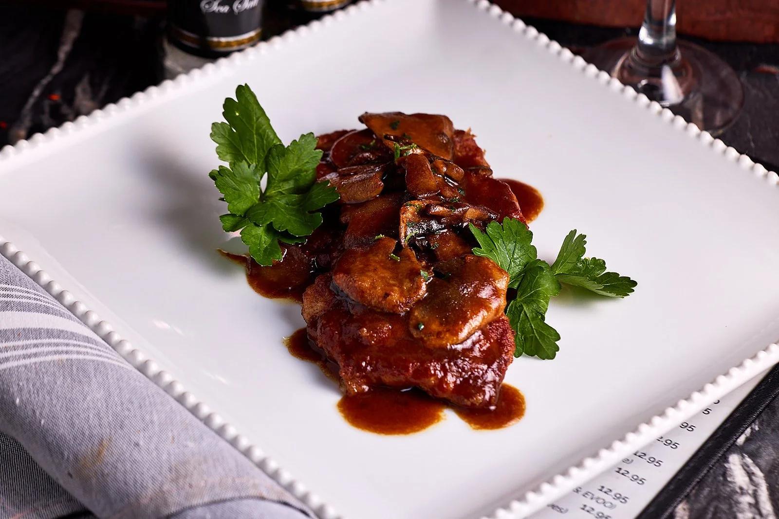 A white square plate with a serving of cooked meat in dark sauce, garnished with fresh parsley leaves, on a black table with wine glasses and a cloth napkin.