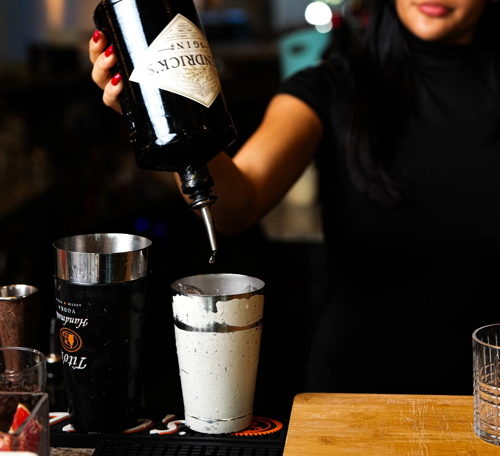Person pouring a cocktail from a bottle into a metal shaker on a bar counter.