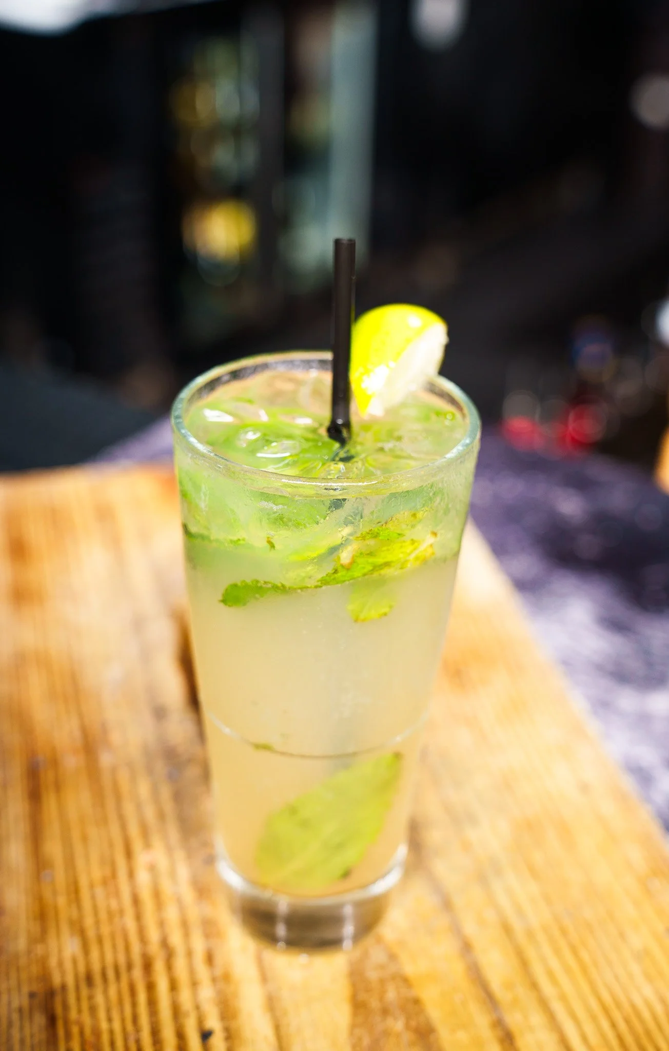 A tall glass of mojito with lime wedge, mint leaves, ice, and a straw on a wooden table.