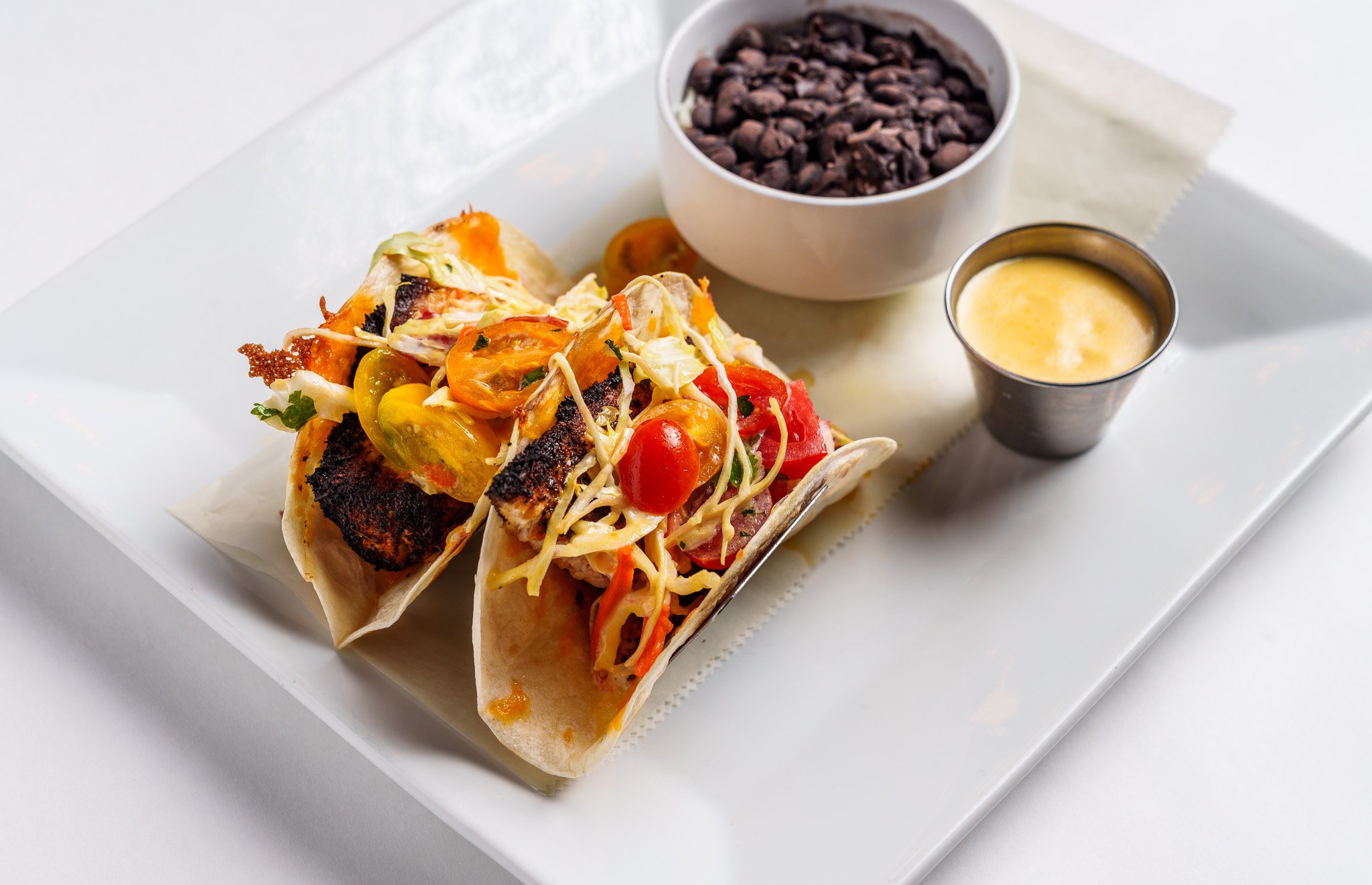 Two tacos filled with grilled fish, cherry tomatoes, and shredded vegetables on a white plate, served with black beans and a cup of yellow sauce.