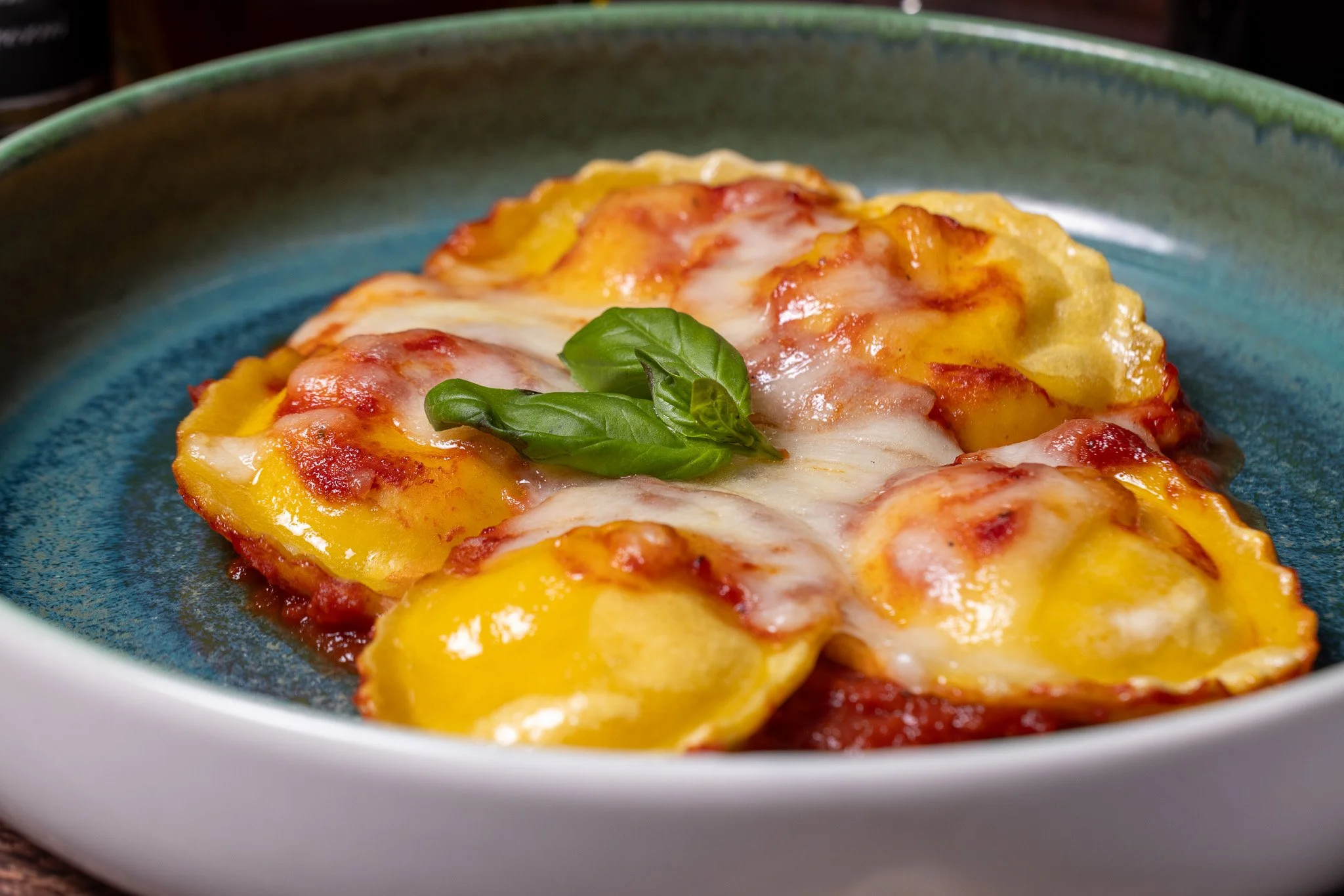 Baked ravioli with melted cheese and fresh basil leaves on top, served in a green dish.