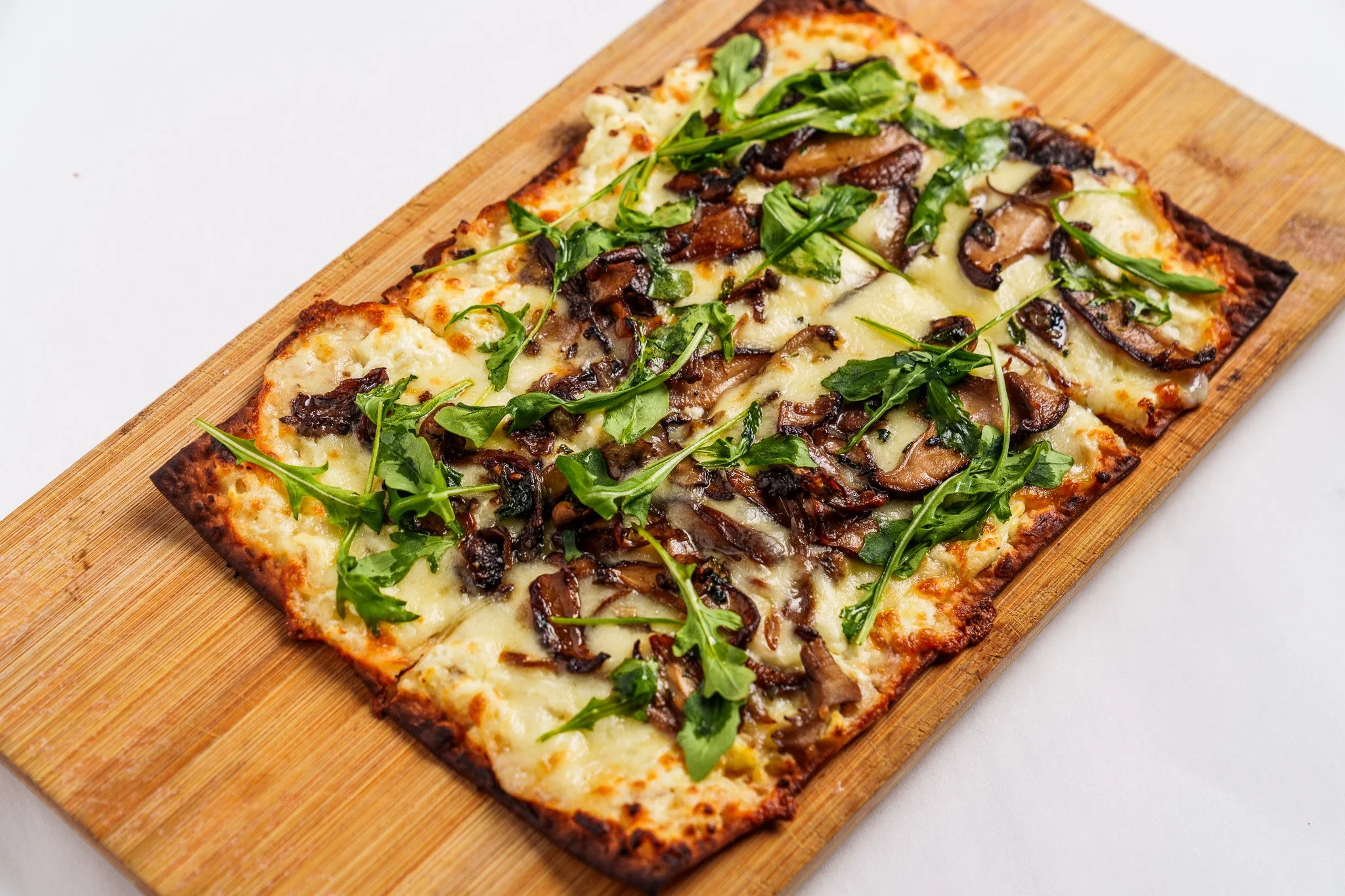A rectangular flatbread topped with melted cheese, sauteed mushrooms, and arugula, on a wooden serving board.