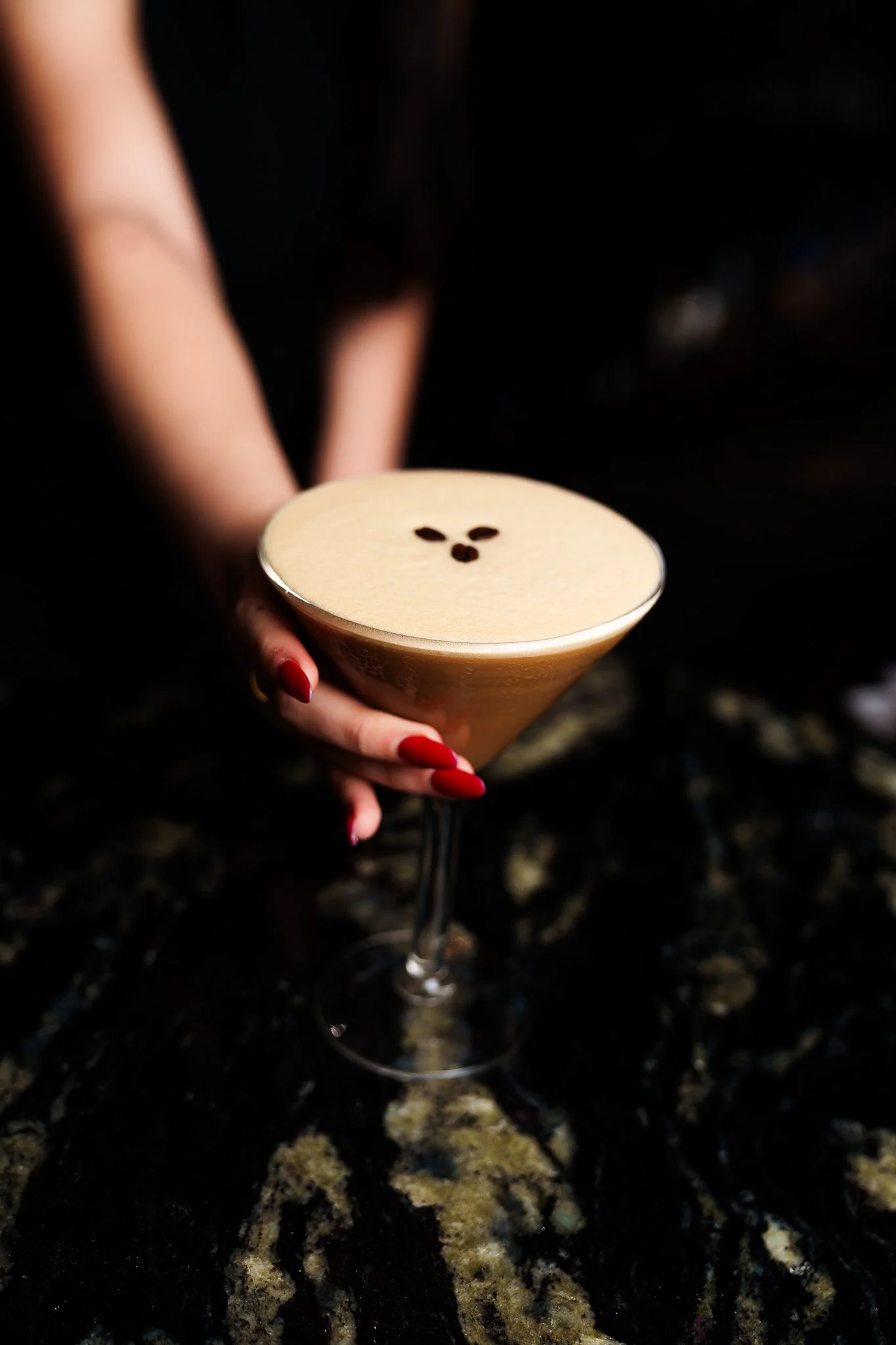 A woman with red nails holding a martini glass filled with a creamy coffee beverage topped with coffee beans arranged to look like a bear's face.