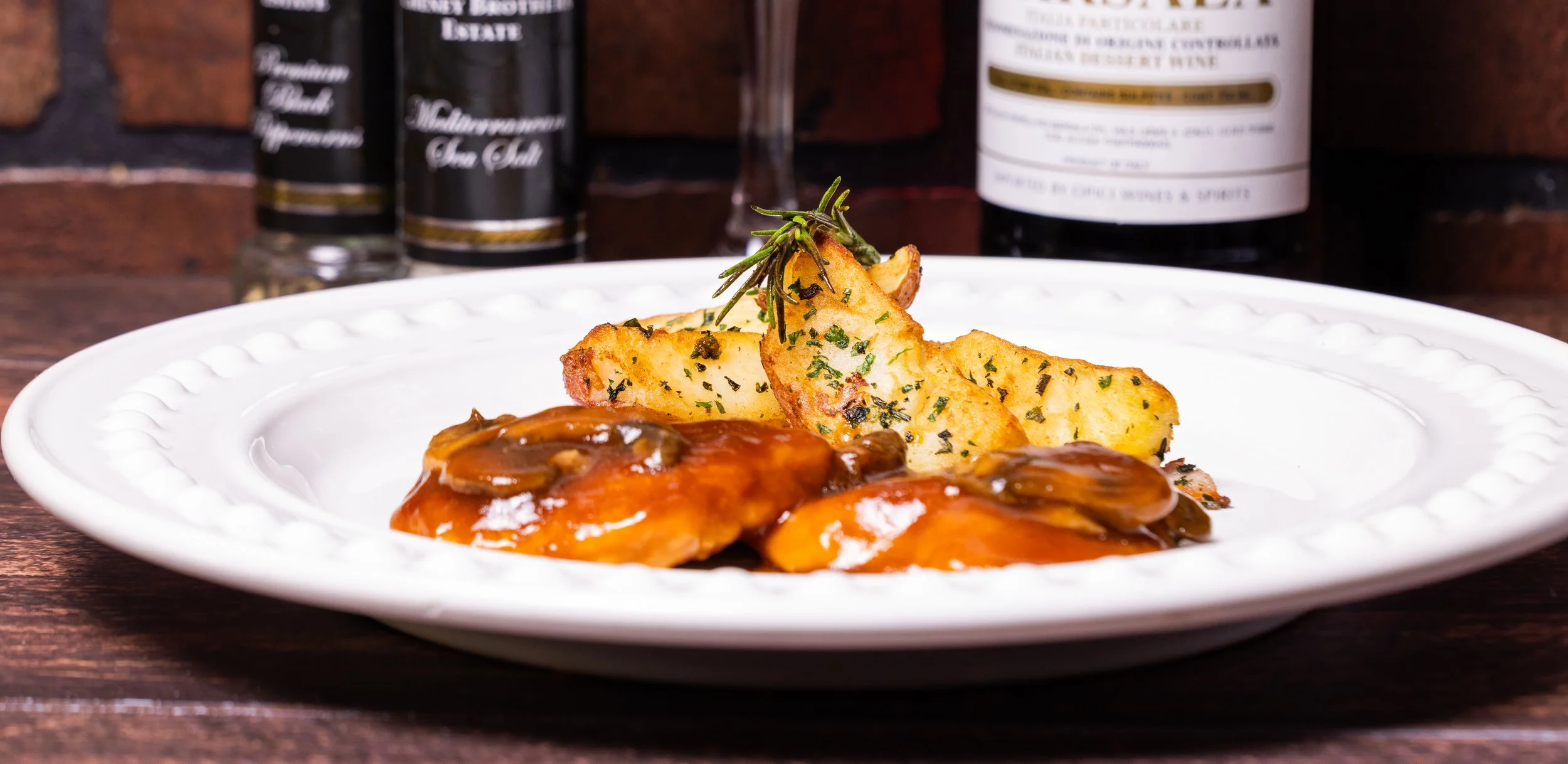 A white plate of roasted chicken with mushrooms, covered in gravy, served with potato wedges garnished with herbs and a sprig of rosemary, on a wooden table with bottles of alcohol in the background.