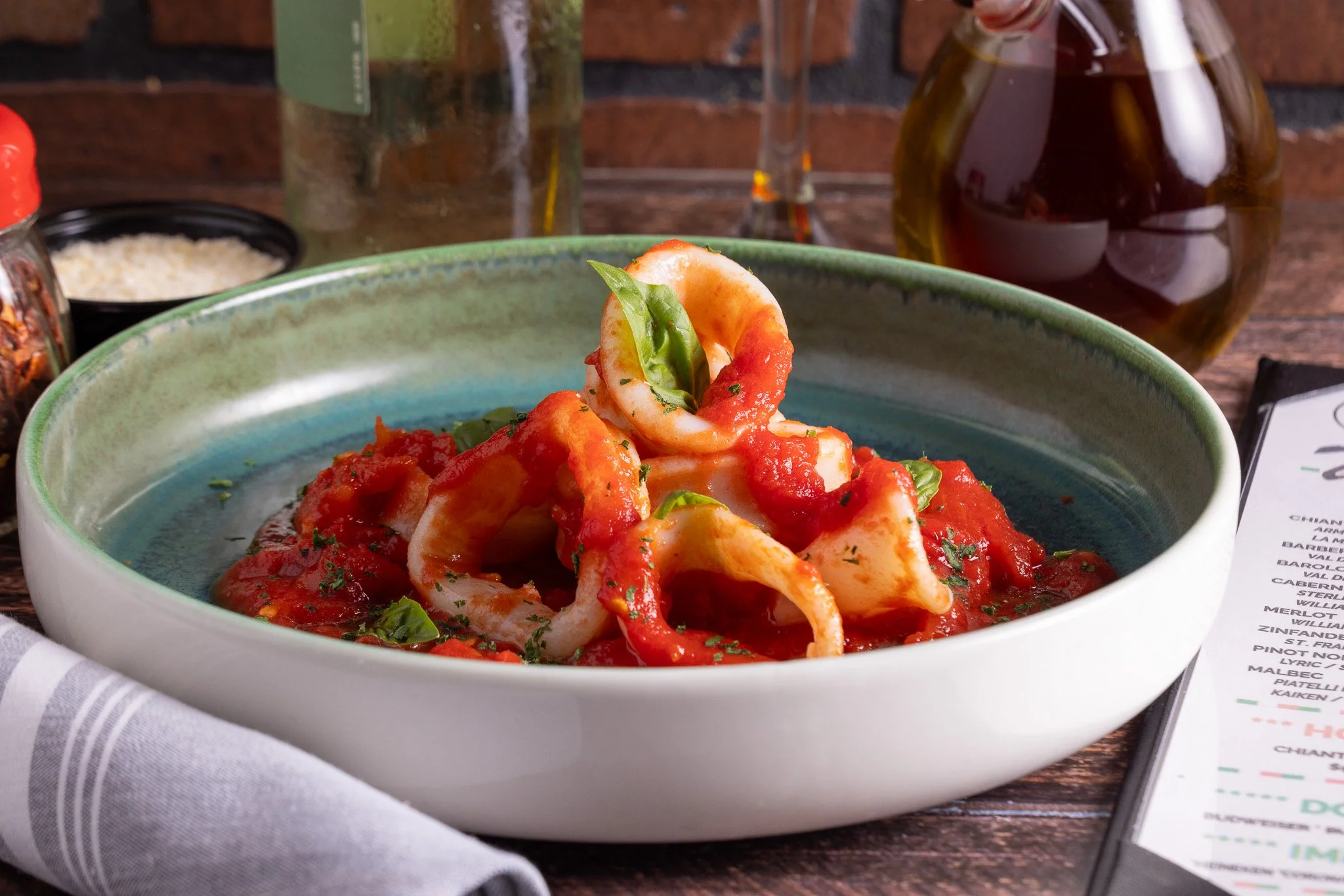 A bowl of pasta with tomato sauce, garnished with basil leaves, on a wooden table with a menu, wine bottles, and condiments in the background.