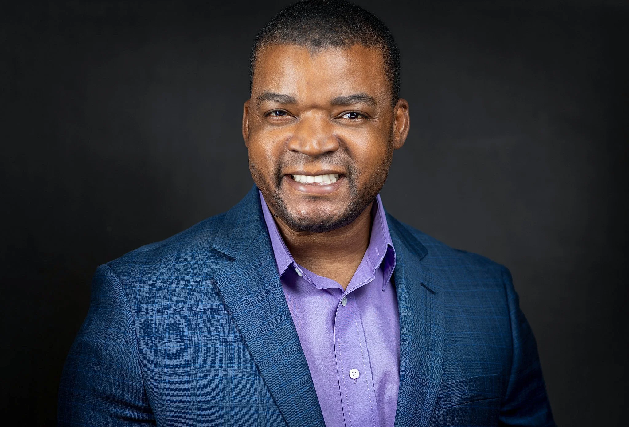 A professional headshot of a smiling Black man with short hair, wearing a blue checked blazer over a lavender dress shirt, against a dark background.