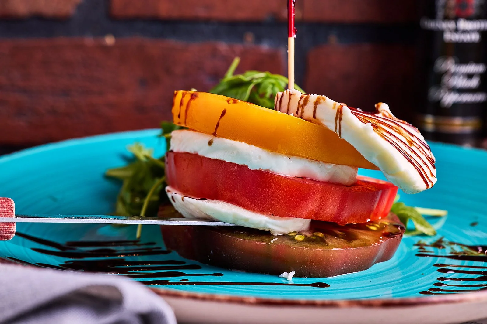 A plate with slices of tomato, mozzarella, yellow and orange vegetables, topped with a drizzle of balsamic glaze and garnished with greens, set on a blue plate.