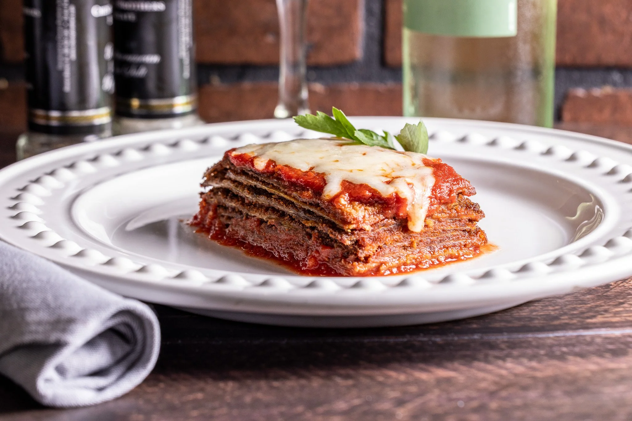 A plate of layered lasagna with tomato sauce, melted cheese on top, garnished with a parsley leaf, on a decorative white plate with a gray napkin nearby, set on a wooden table with a brick wall background.
