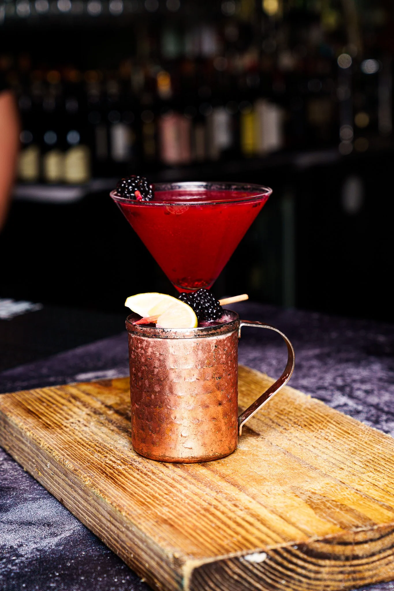 A copper mug and a martini glass with blackberries and lemon wedge on a wooden serving board, set on a dark bar counter.
