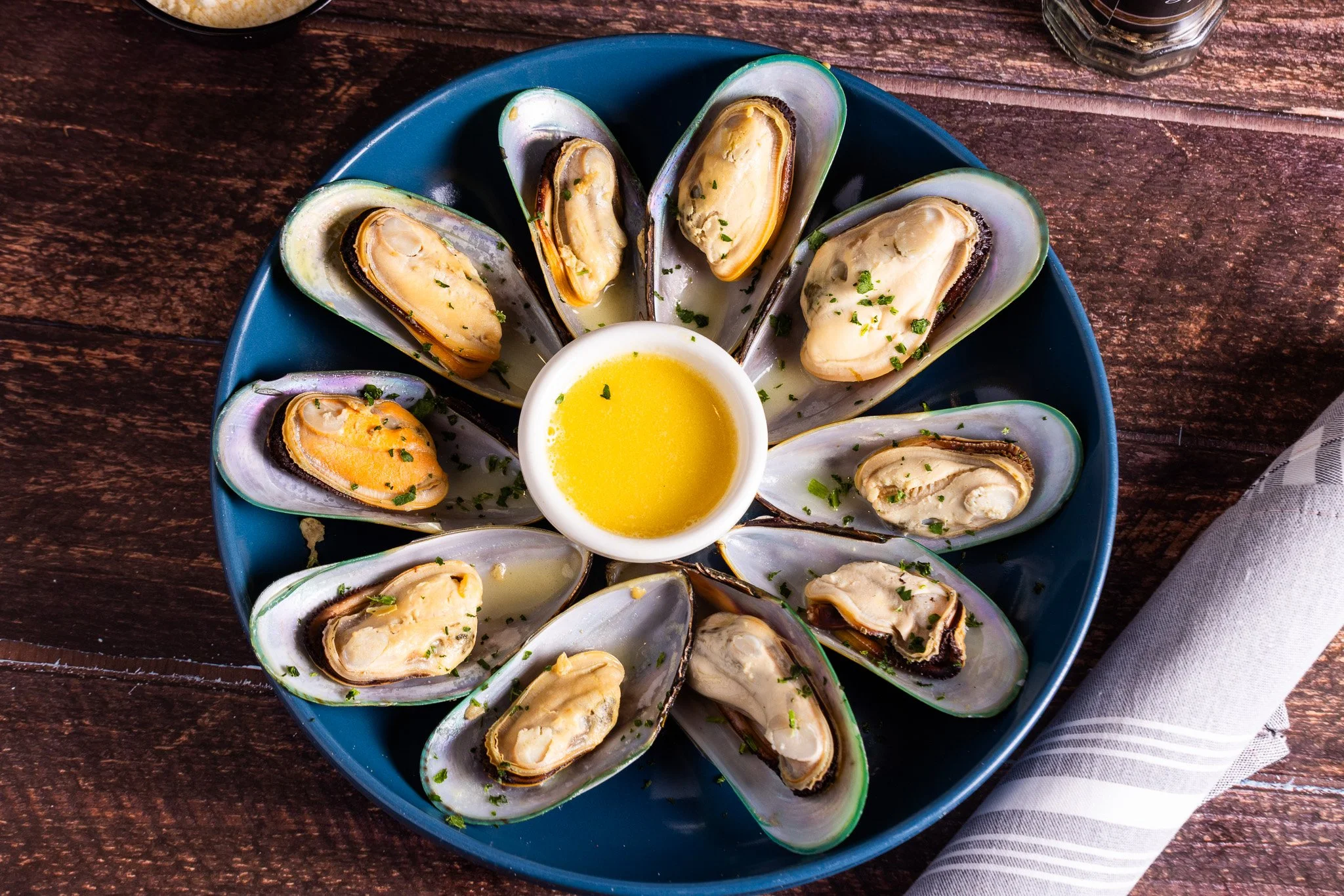 A blue plate of cooked mussels garnished with herbs, served with a small bowl of melted butter in the center on a wooden table.