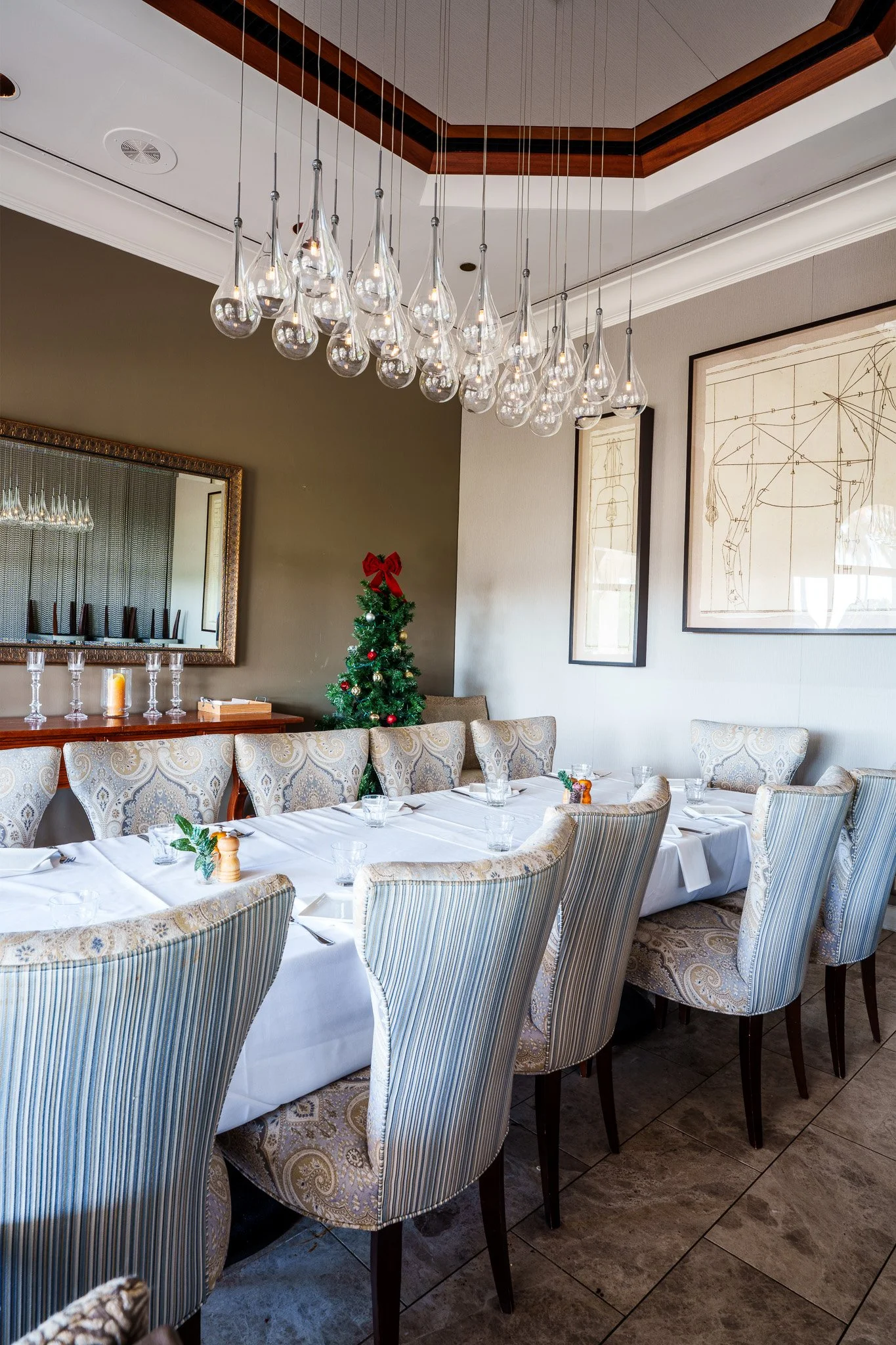 Elegant dining room decorated for Christmas with a small Christmas tree, table set with glasses and silverware, and a modern chandelier hanging from the ceiling.