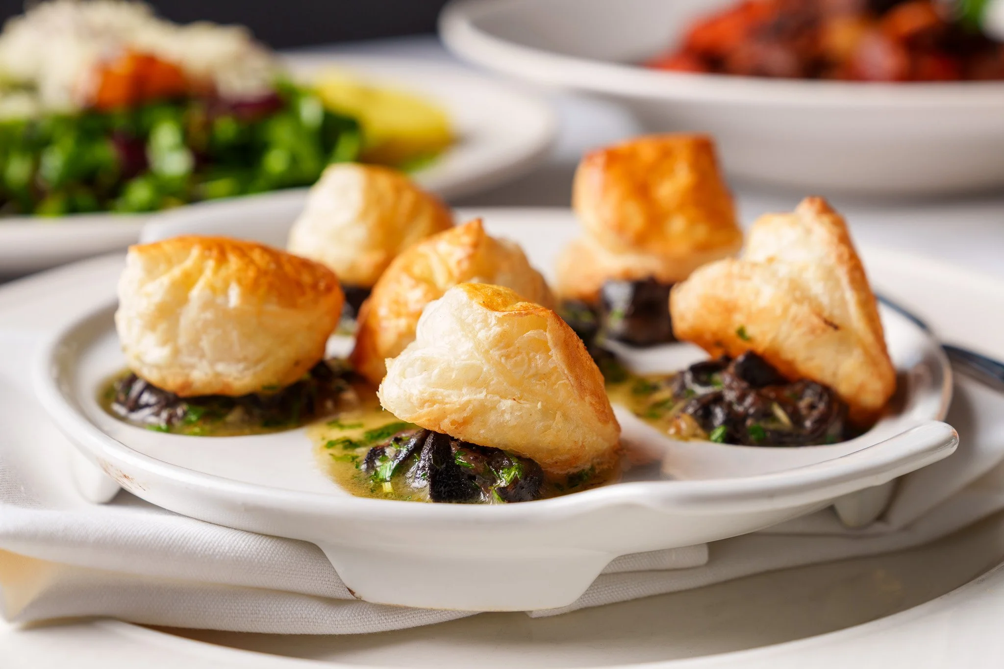 Close-up of a dish with small savory puff pastry appetizers placed on a white plate, with green herbs and a brown sauce underneath, and blurred salads in the background.